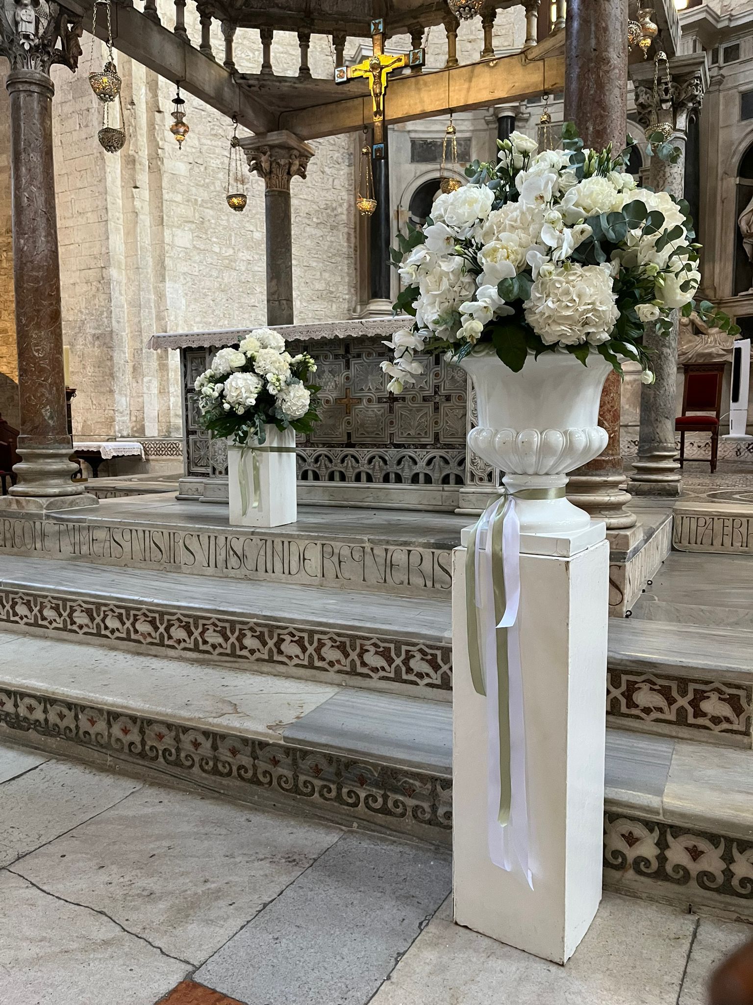 Composizioni floreali bianche in urne su piedistalli all'interno di una chiesa.