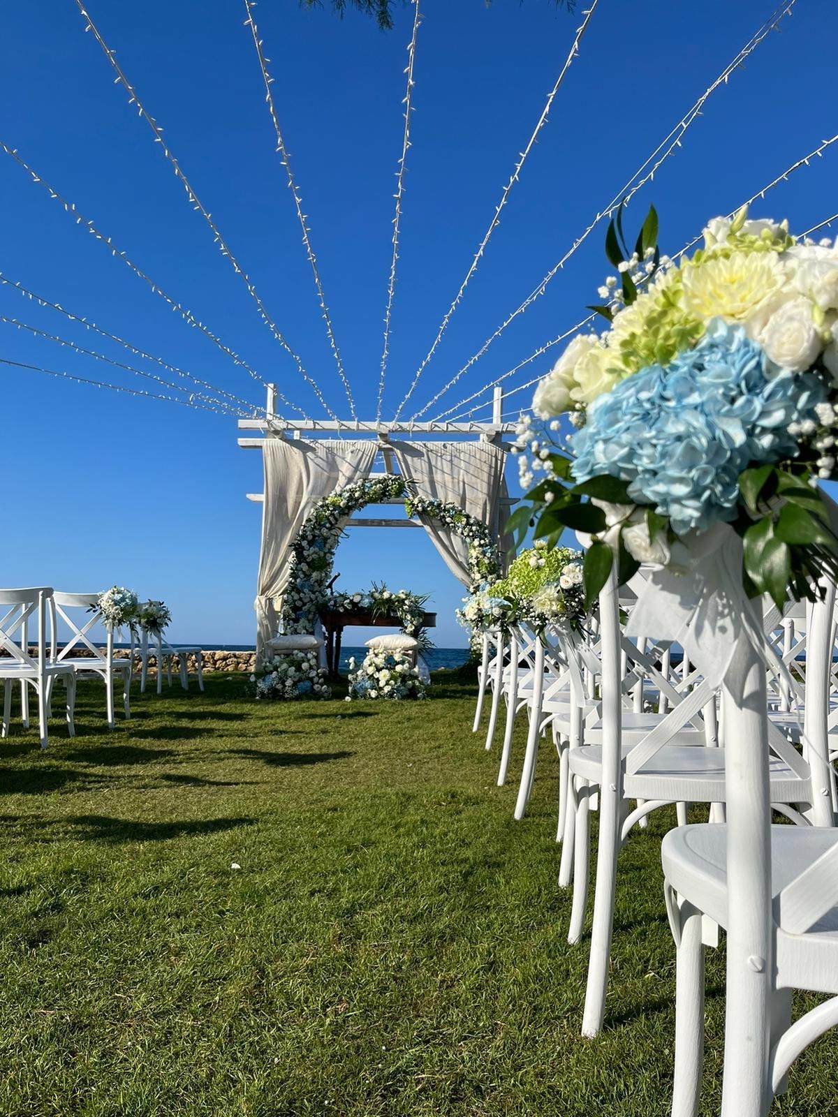 Allestimento per matrimonio su un prato verde con sedie bianche, composizioni floreali e un arco decorato sotto un cielo azzurro.