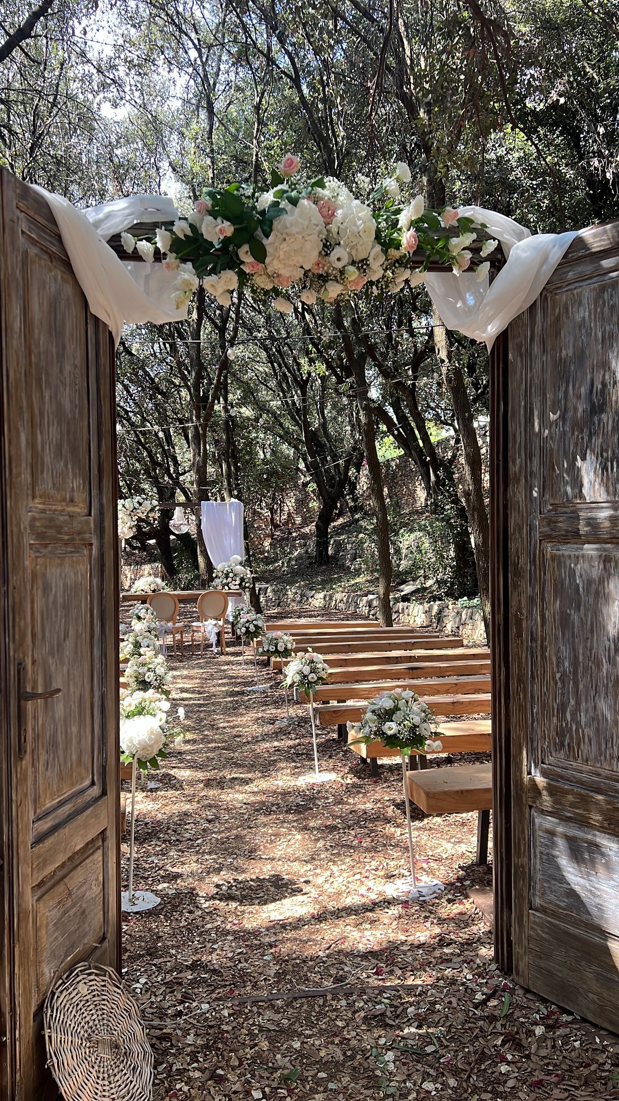 Porte di legno si aprono su una cerimonia nuziale nel bosco. Un arco floreale decora l'ingresso. Panche di legno sono allestite per gli ospiti.