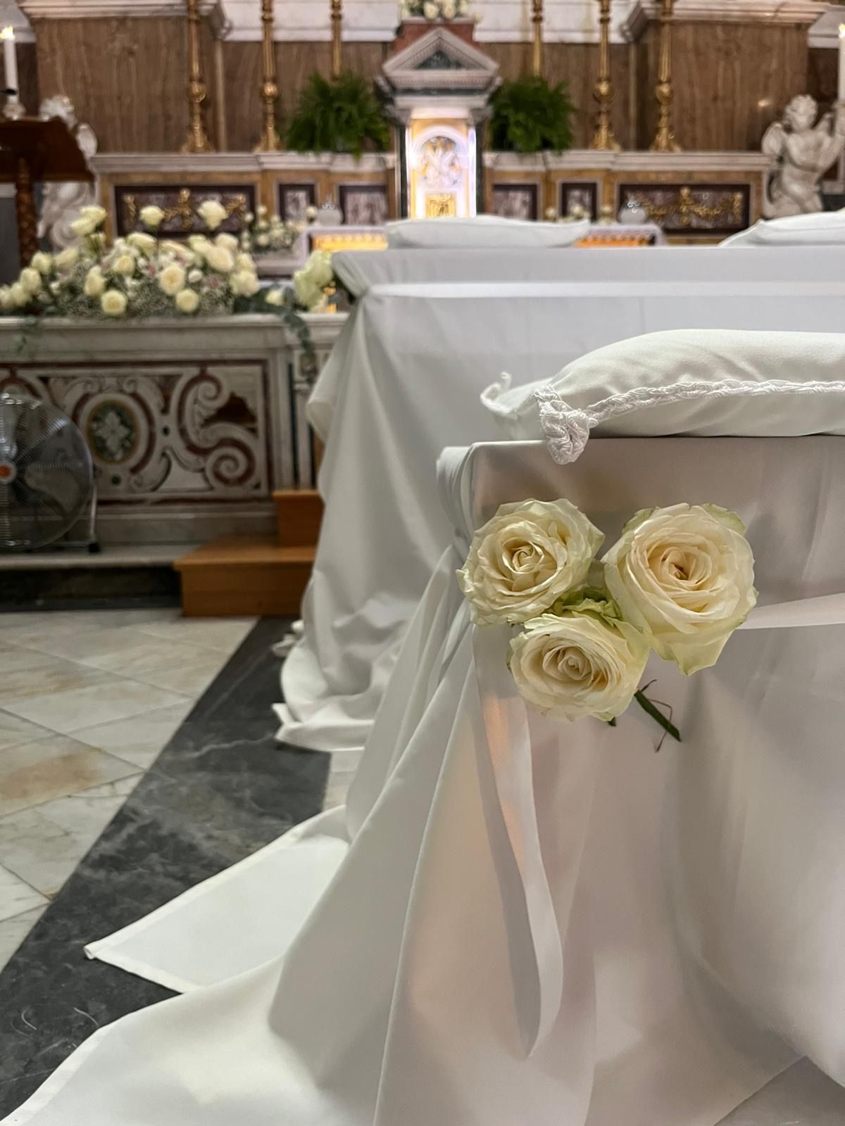 Tavoli con drappeggi bianchi in una chiesa, decorati con rose bianche e cuscini. Sullo sfondo un altare e composizioni floreali.