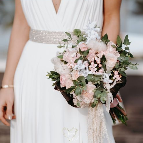 Sposa in abito bianco, con in mano un bouquet di fiori rosa e verdi e una cintura tempestata di gioielli.