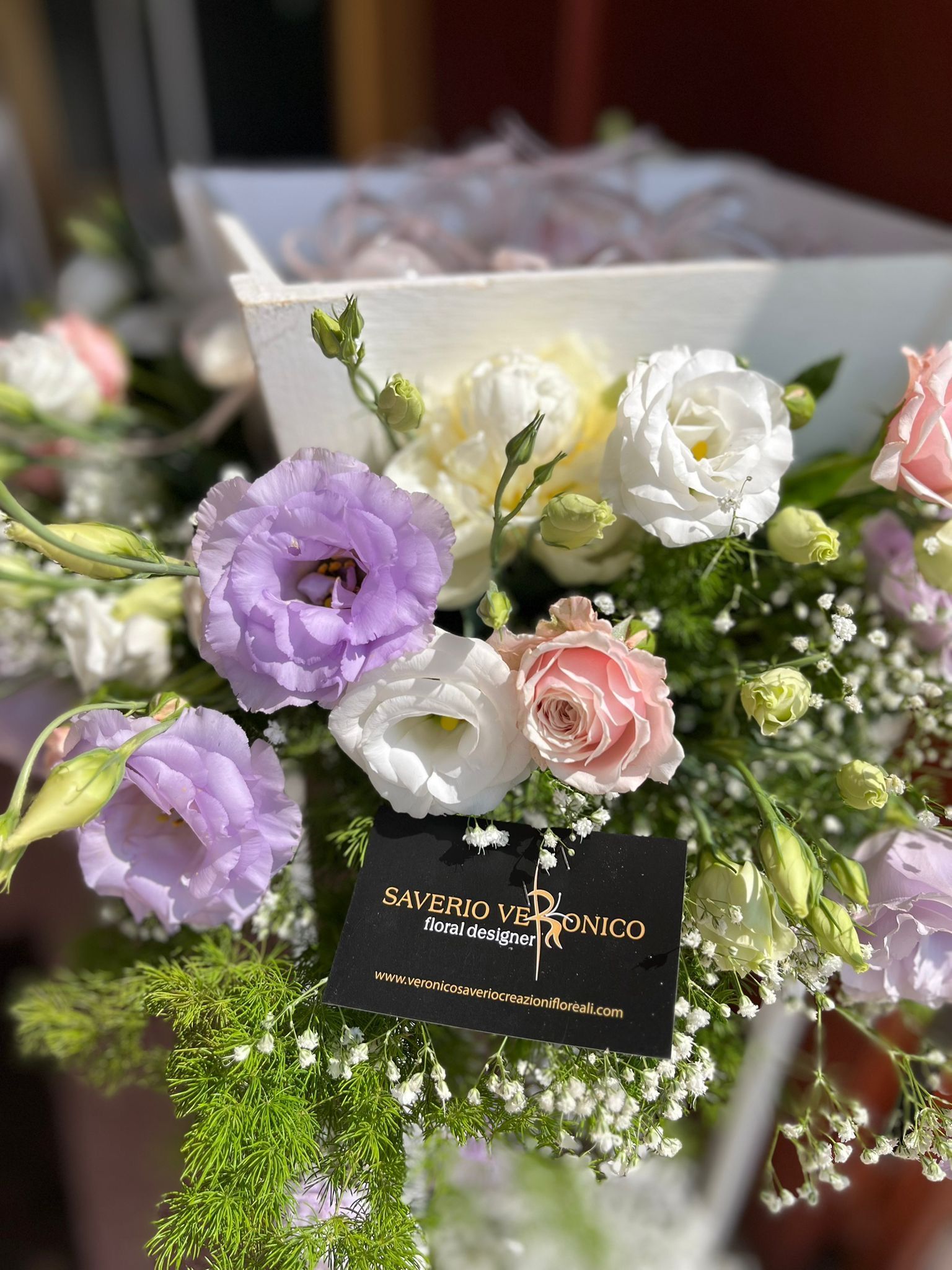 Primo piano di una composizione floreale in un contenitore bianco, con fiori viola, bianchi e rosa, verde e un cartoncino nero.