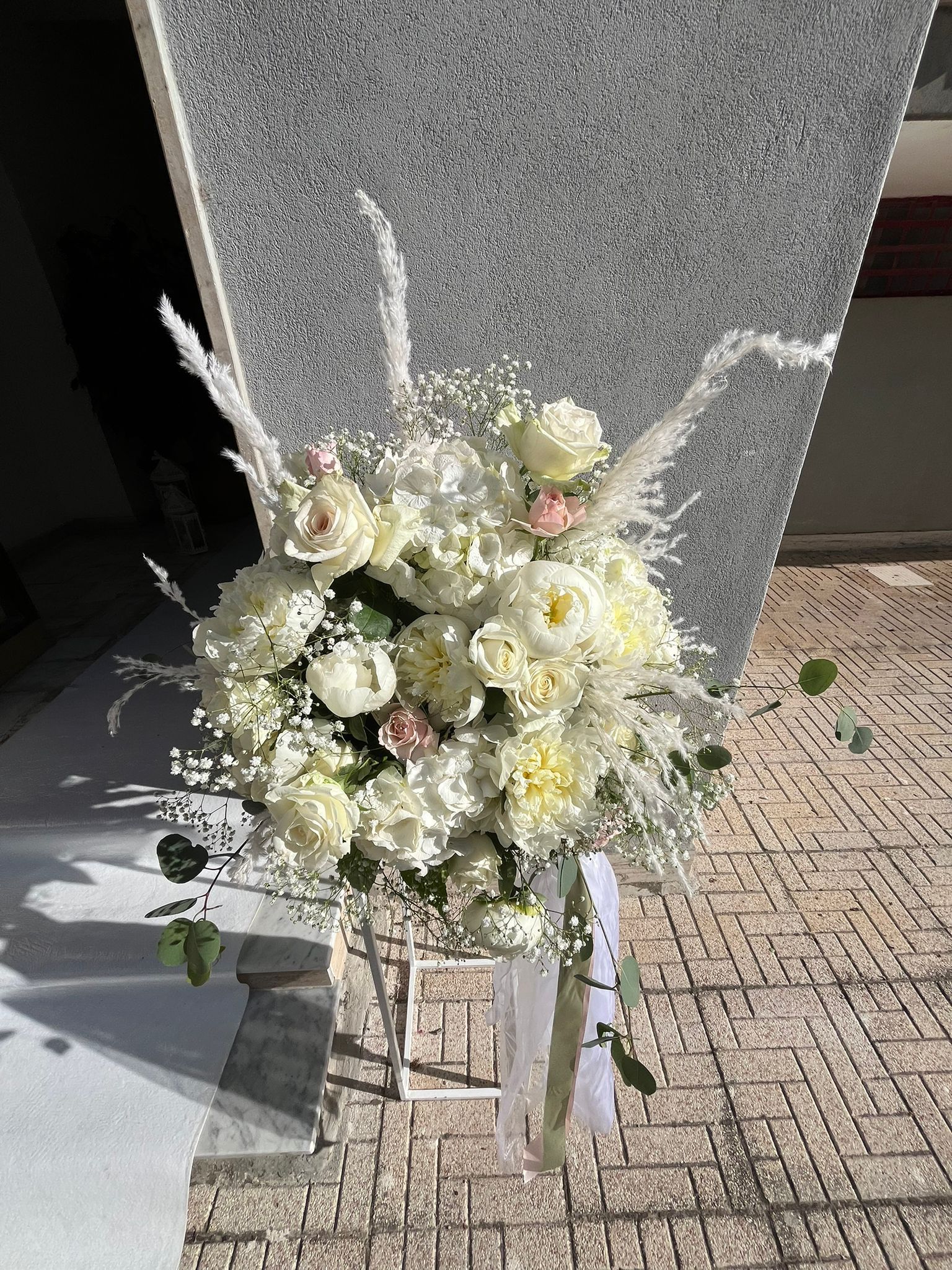 Composizione floreale bianca su un supporto bianco, contro un muro di pietra grigia e un pavimento in mattoni.