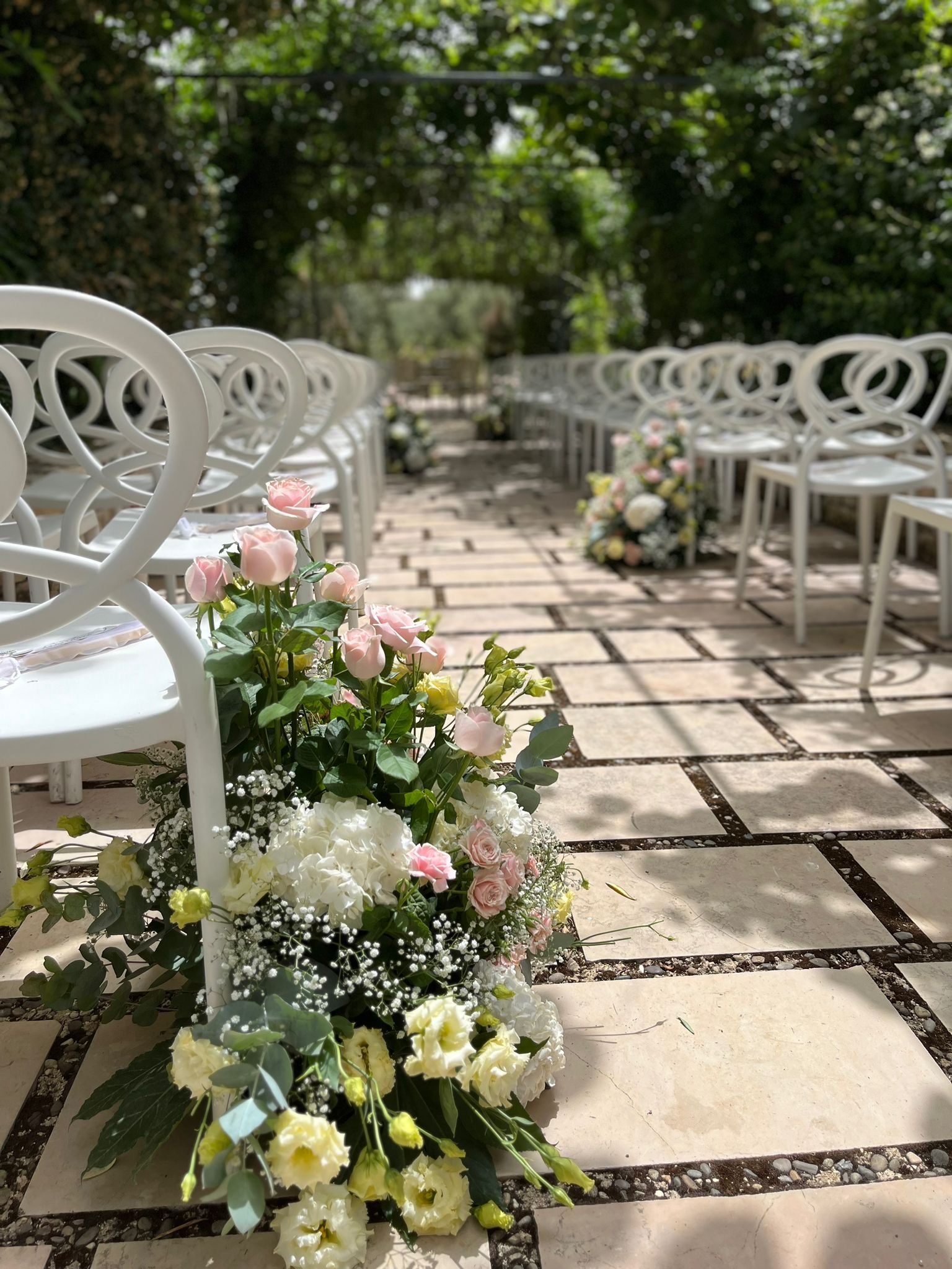 Allestimento della cerimonia nuziale: sedie bianche costeggiano un sentiero in pietra, decorato con composizioni floreali.