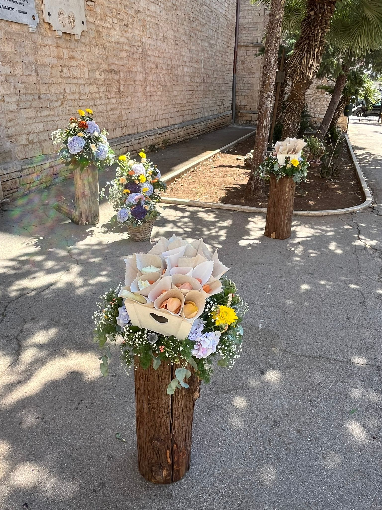 Composizioni floreali su rustici ceppi di legno su una superficie pavimentata vicino a un muro di mattoni e ad alcuni alberi.