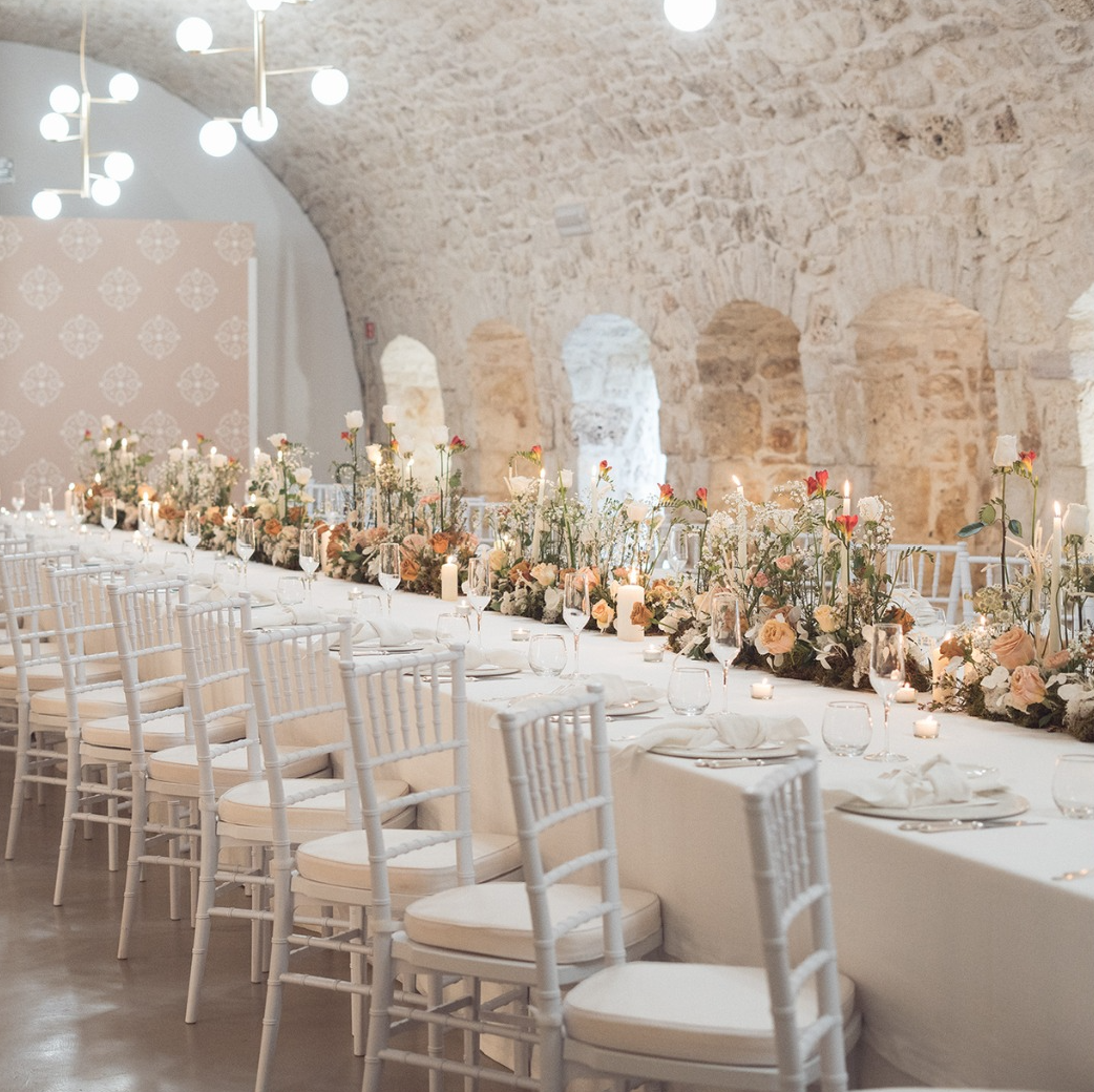 Lungo tavolo da pranzo all'aperto apparecchiato per una festa, decorato con fiori e candele.