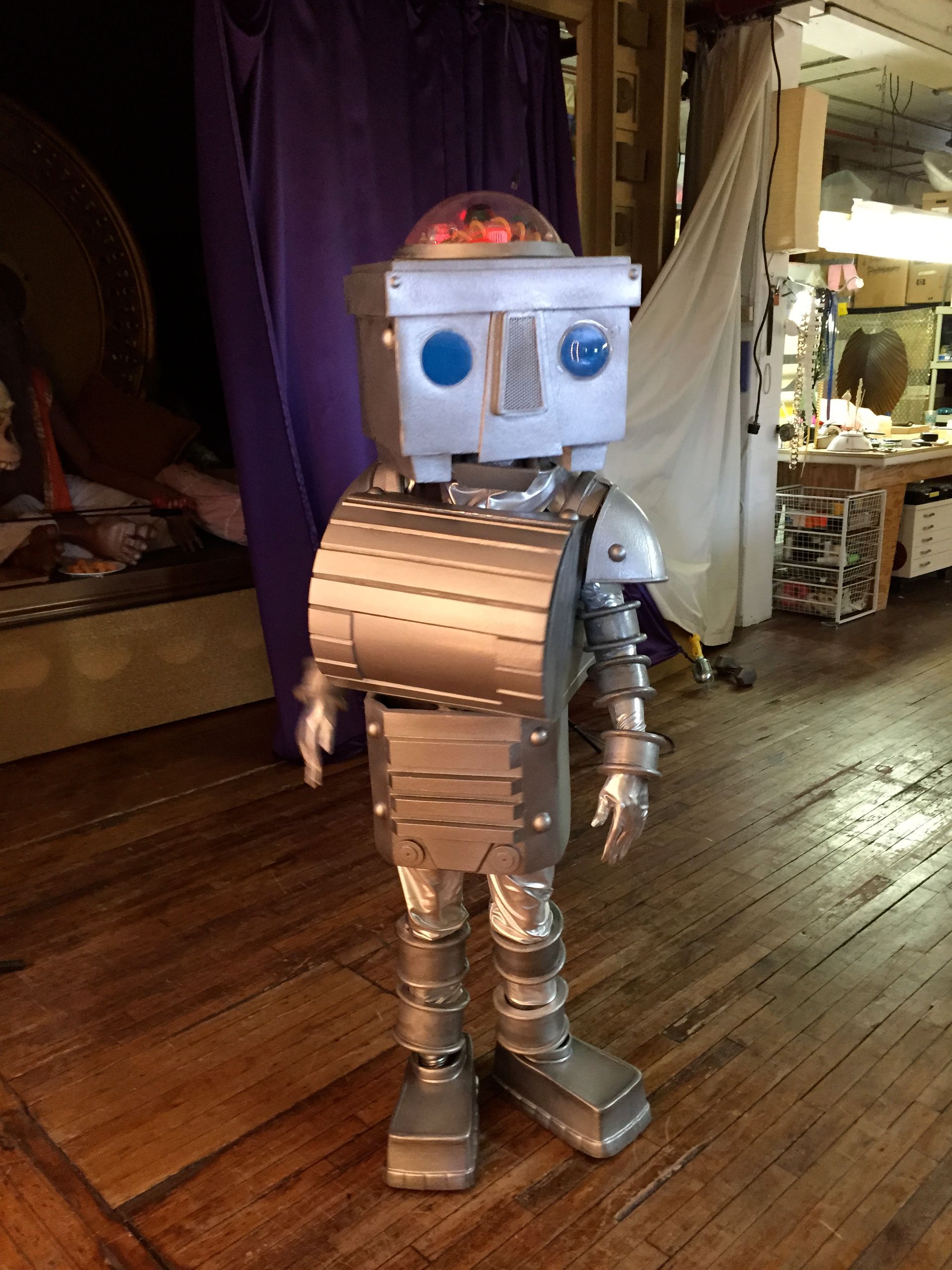 A robot is standing on a wooden floor in front of a purple curtain.