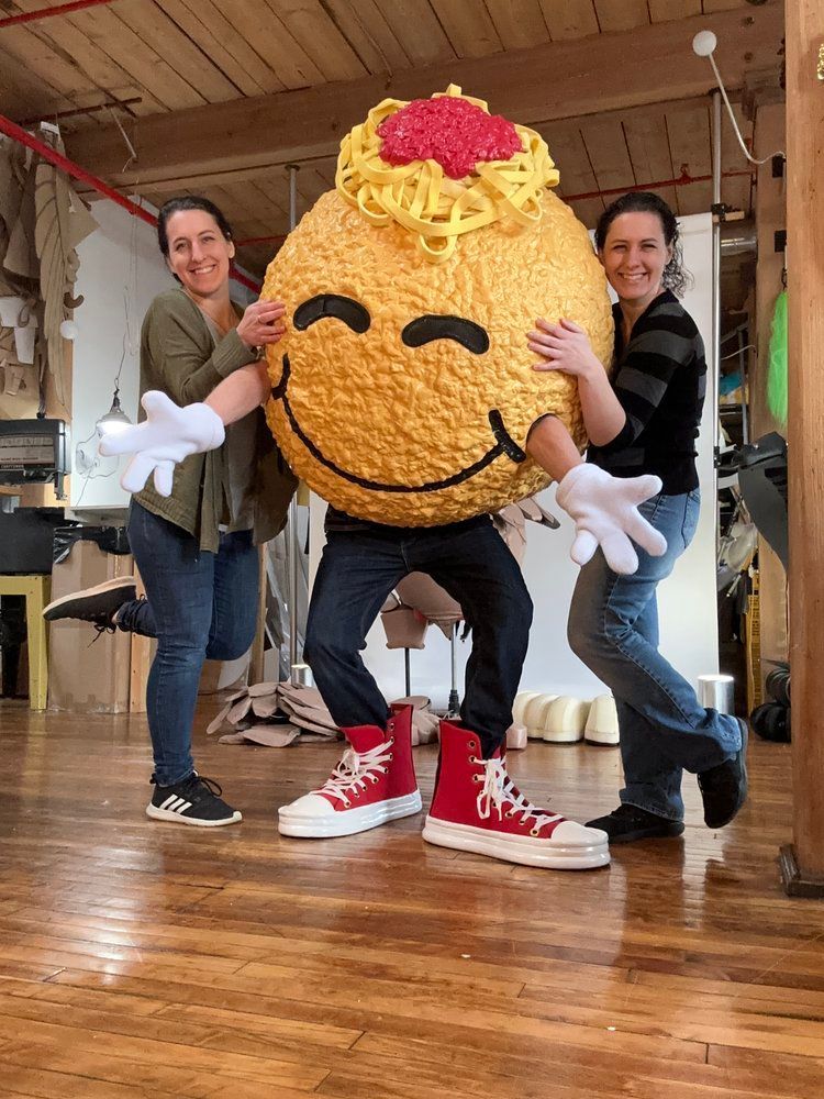 Two women are standing next to a giant smiley face mascot.