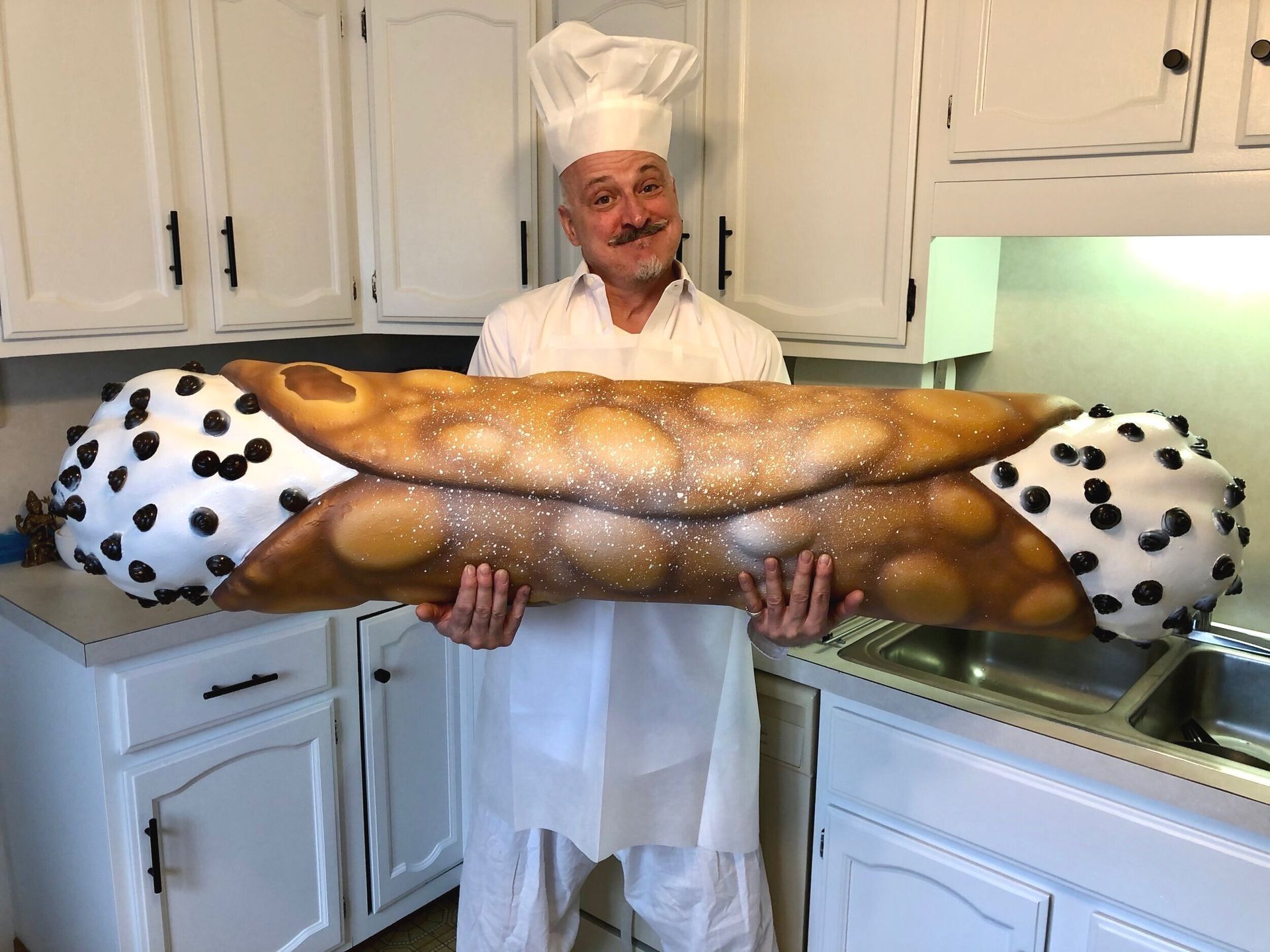 A man in a chef 's hat is holding a giant cannoli in a kitchen.