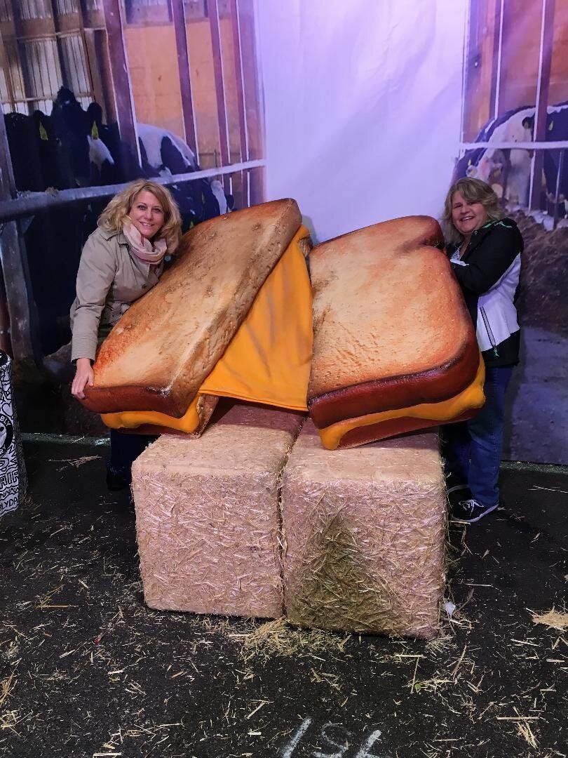 Two women are standing next to a giant sandwich made out of hay.