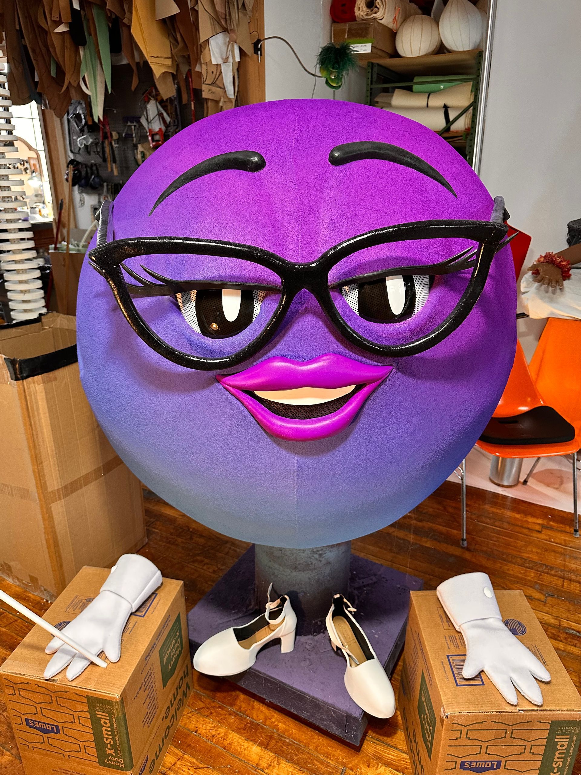 Purple-faced emoji wearing glasses, with gloves and shoes on boxes, and a craft setting.