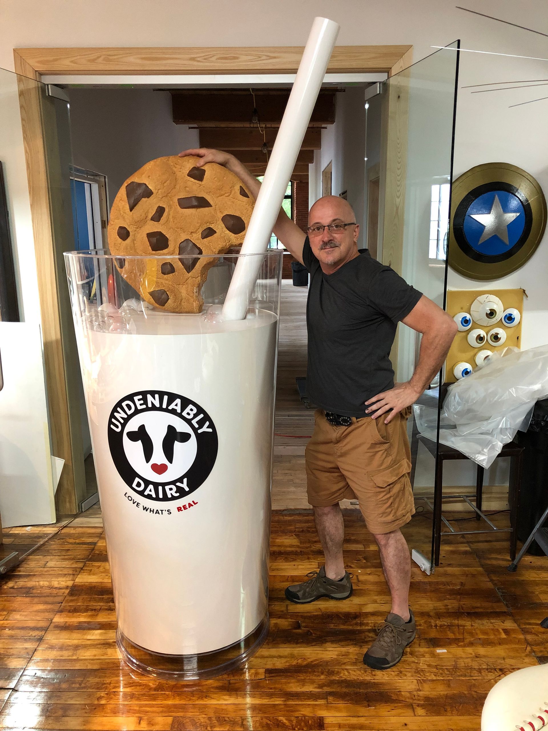 Man poses with giant milk carton costume with a cookie and straw; indoor setting.