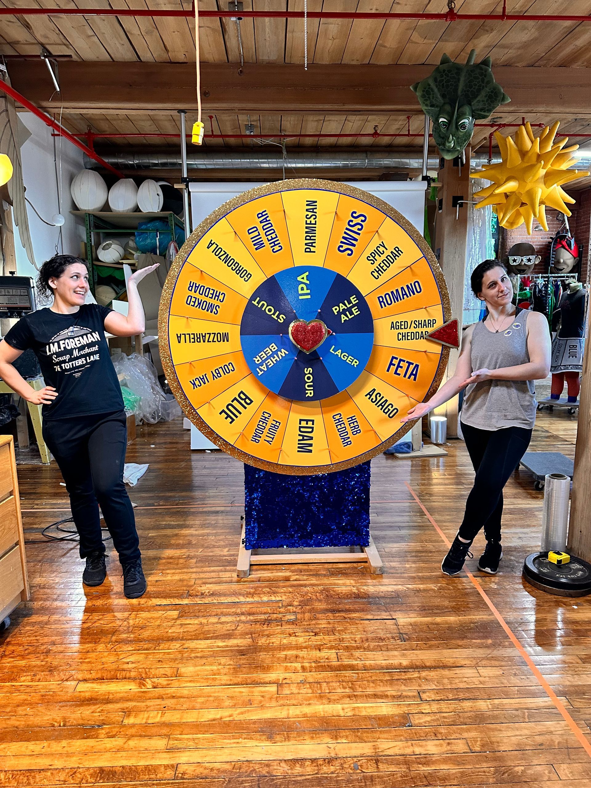 Two women pose with a large prize wheel, celebrating in a colorful room.