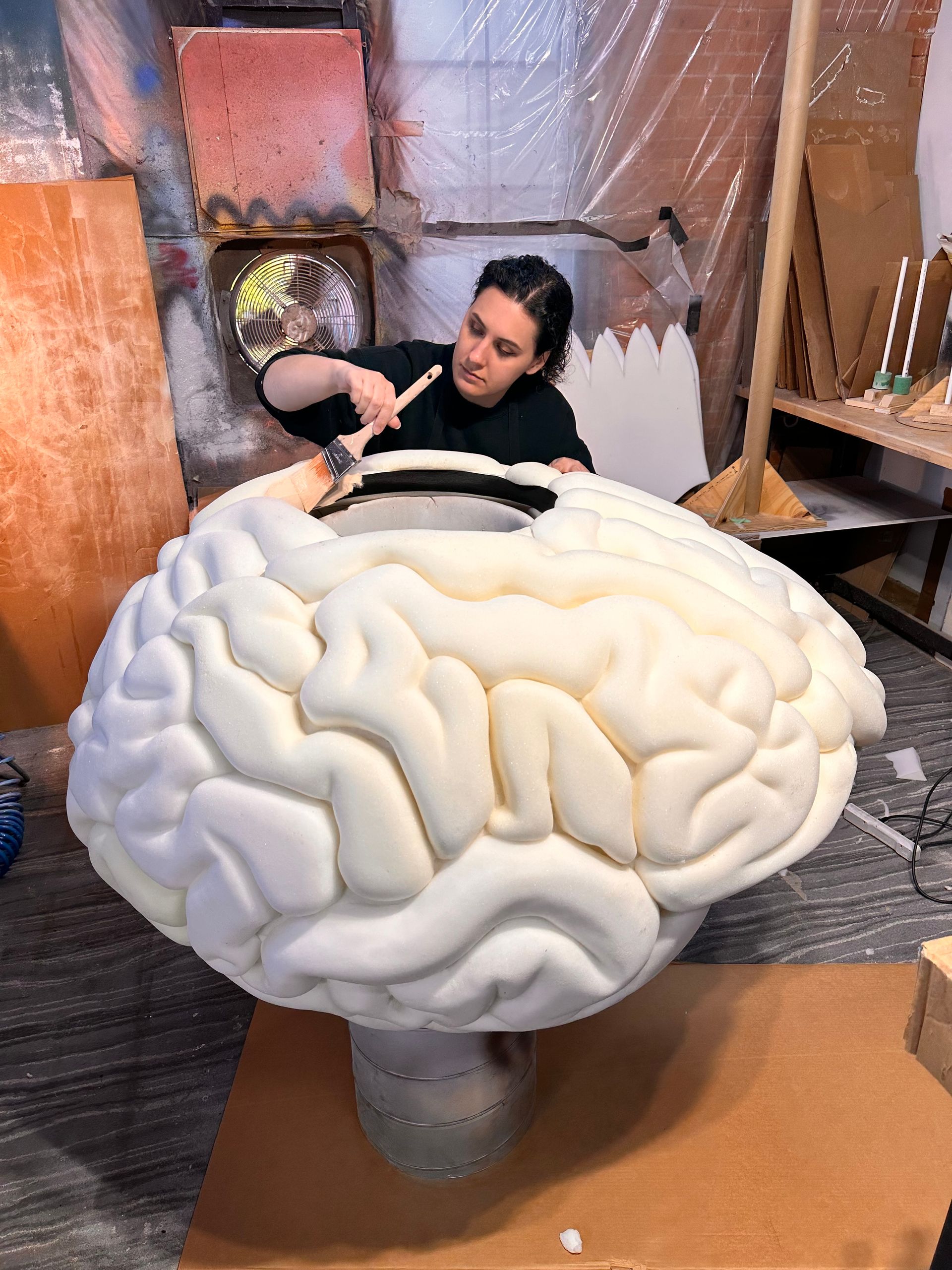 Woman painting on a large foam brain sculpture in a workshop.