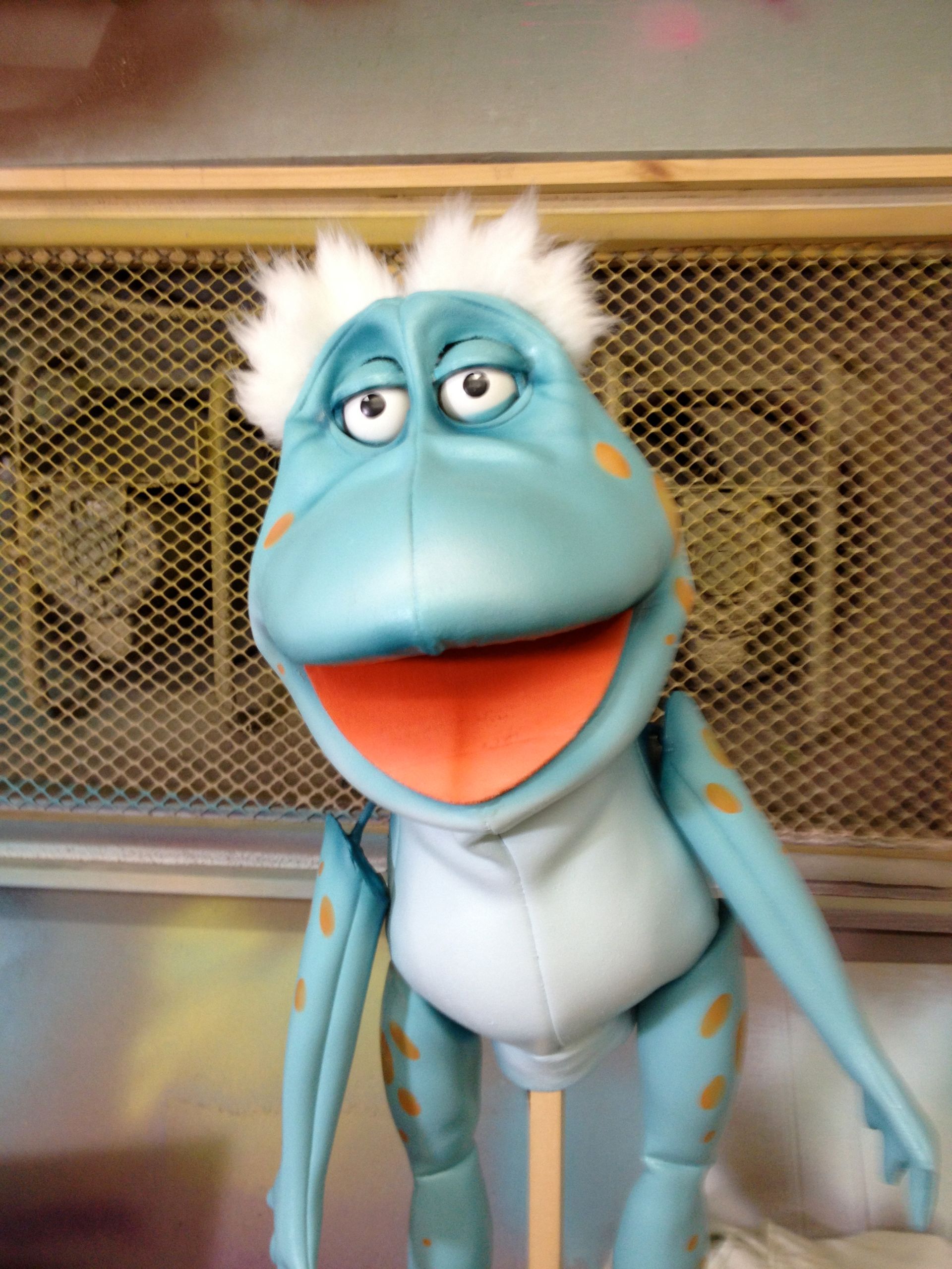 A blue frog puppet with white hair is standing on a stick