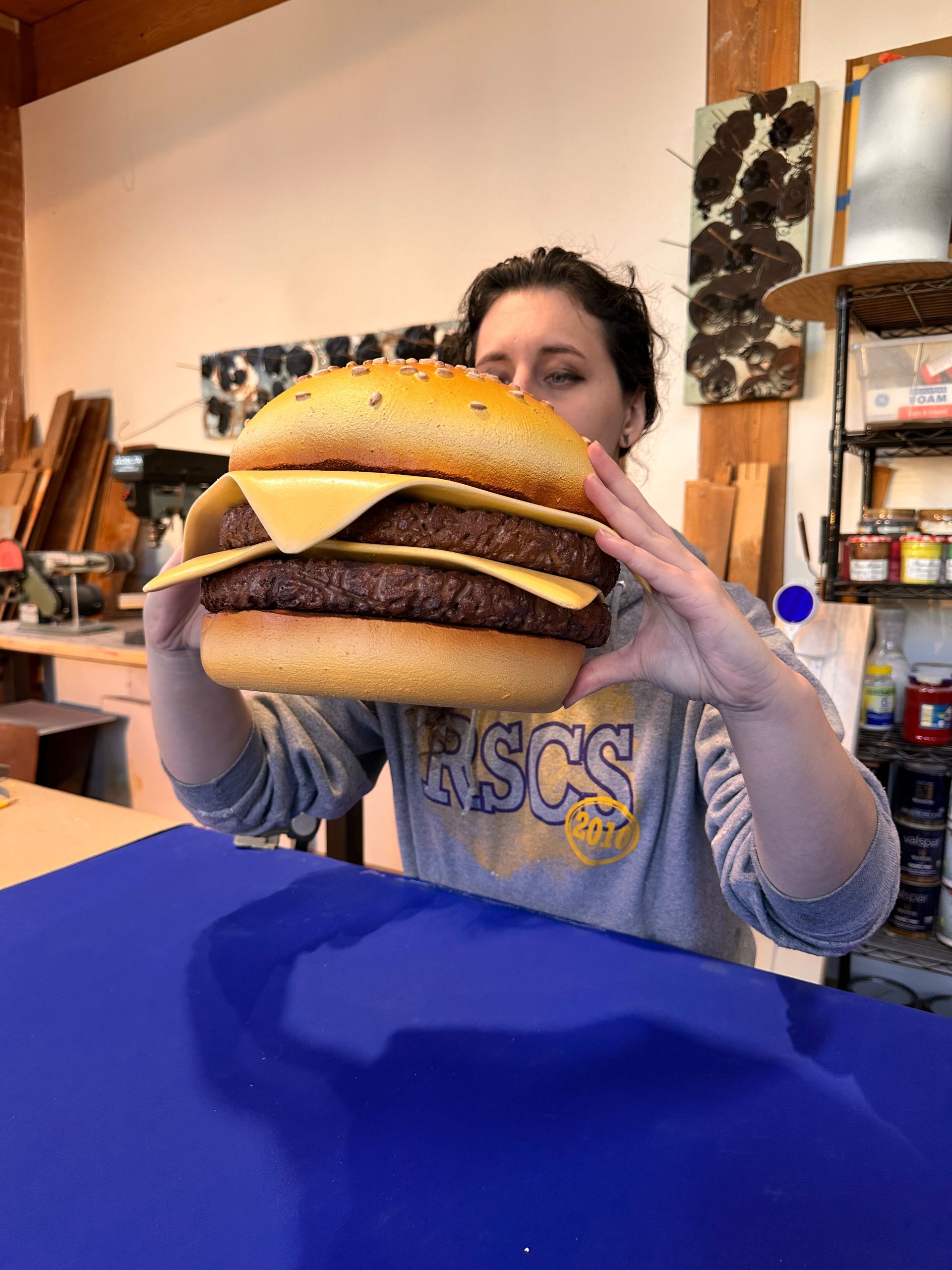 Woman holding a large burger in a workshop, smiling. Blue table in the foreground.
