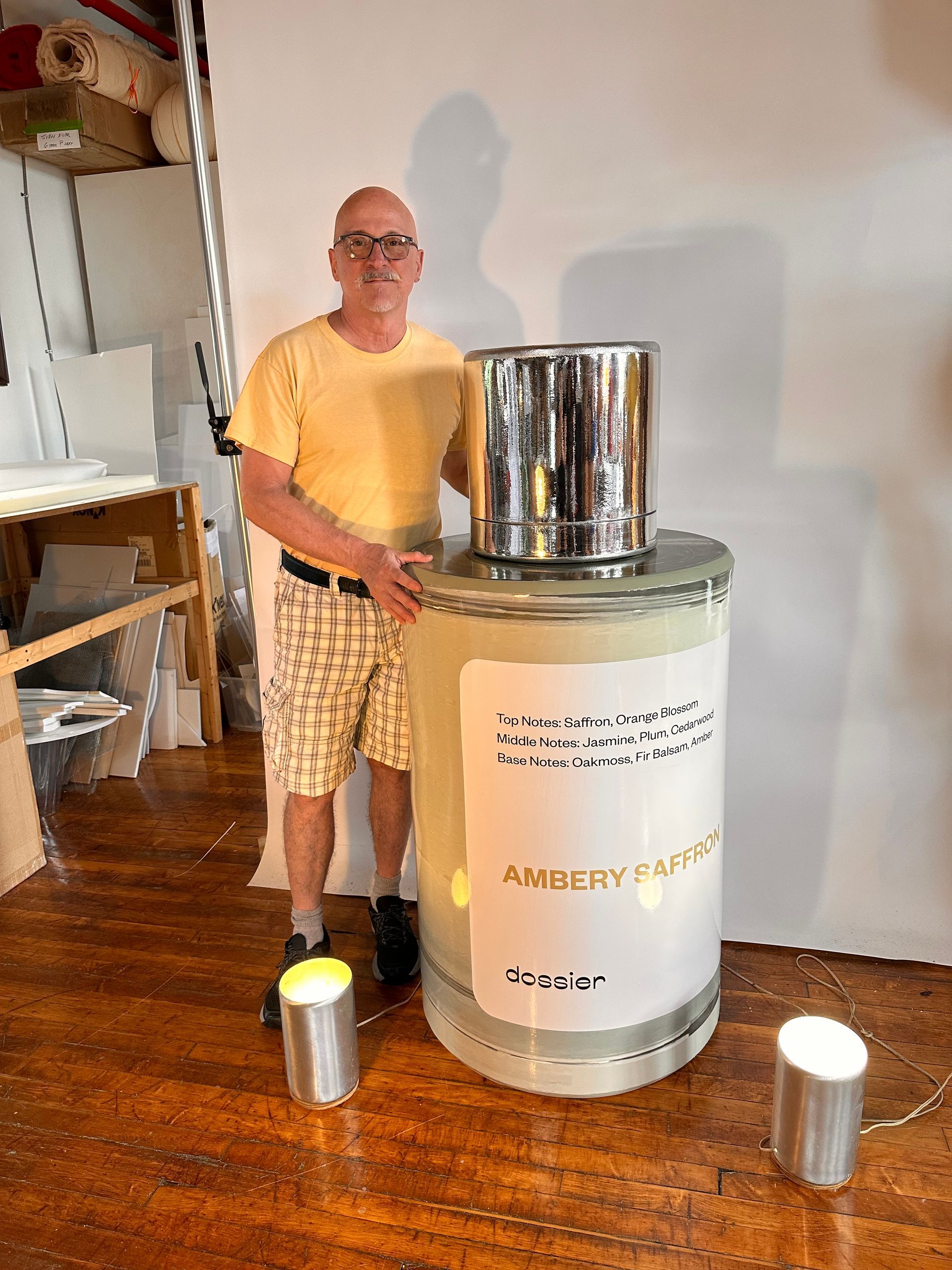 Man stands beside a giant perfume bottle. He's in shorts and a yellow shirt. Indoors, with small lamps.