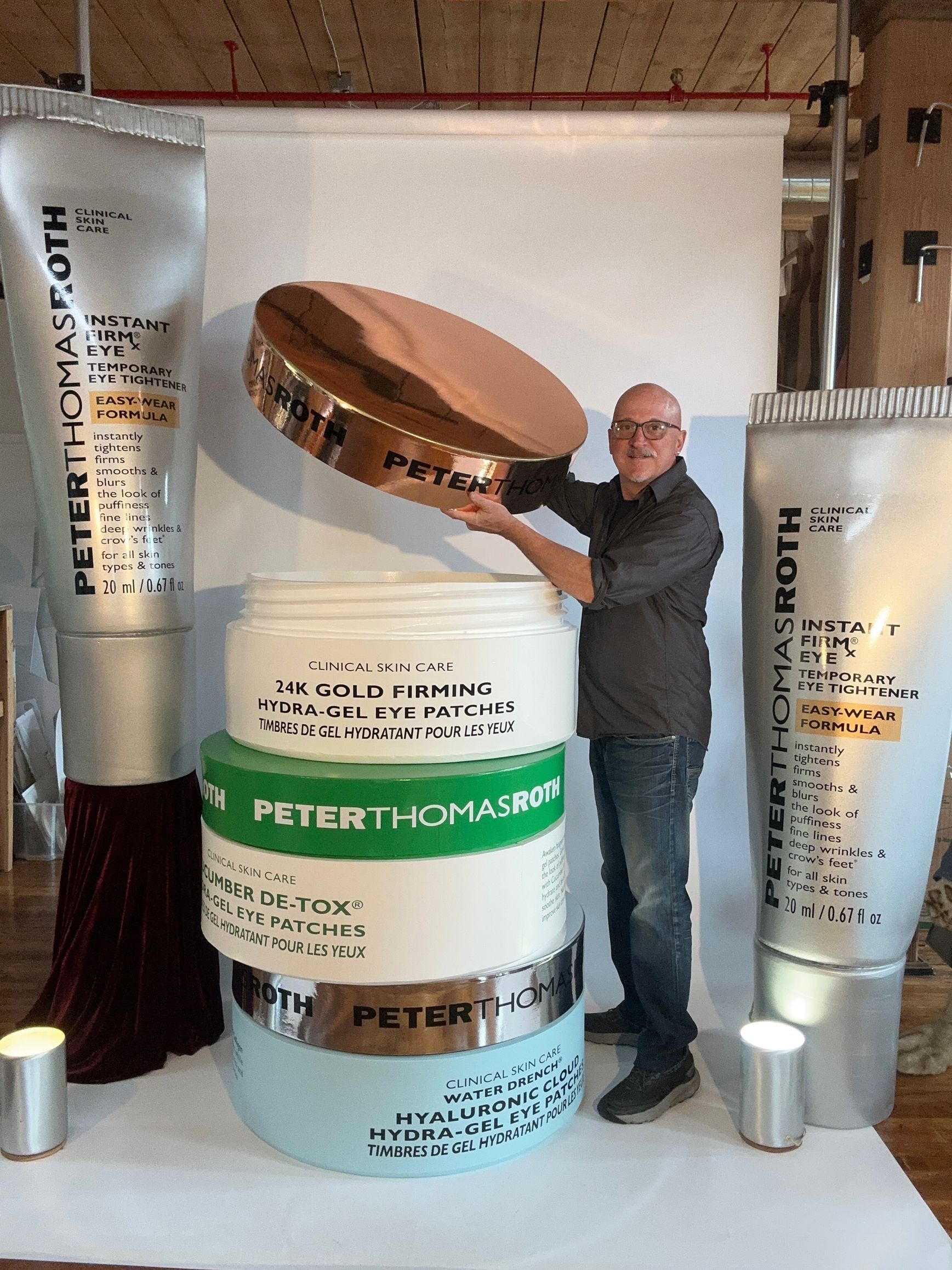 Man holding giant skincare jar lid, standing by oversized Peter Thomas Roth products. Studio setting.