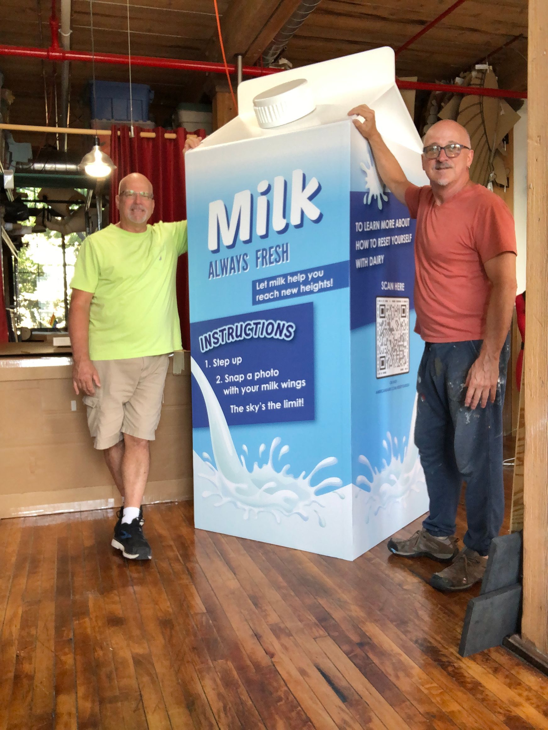 Two men stand next to a large cardboard milk carton prop; one wears a lime shirt, other an orange shirt.
