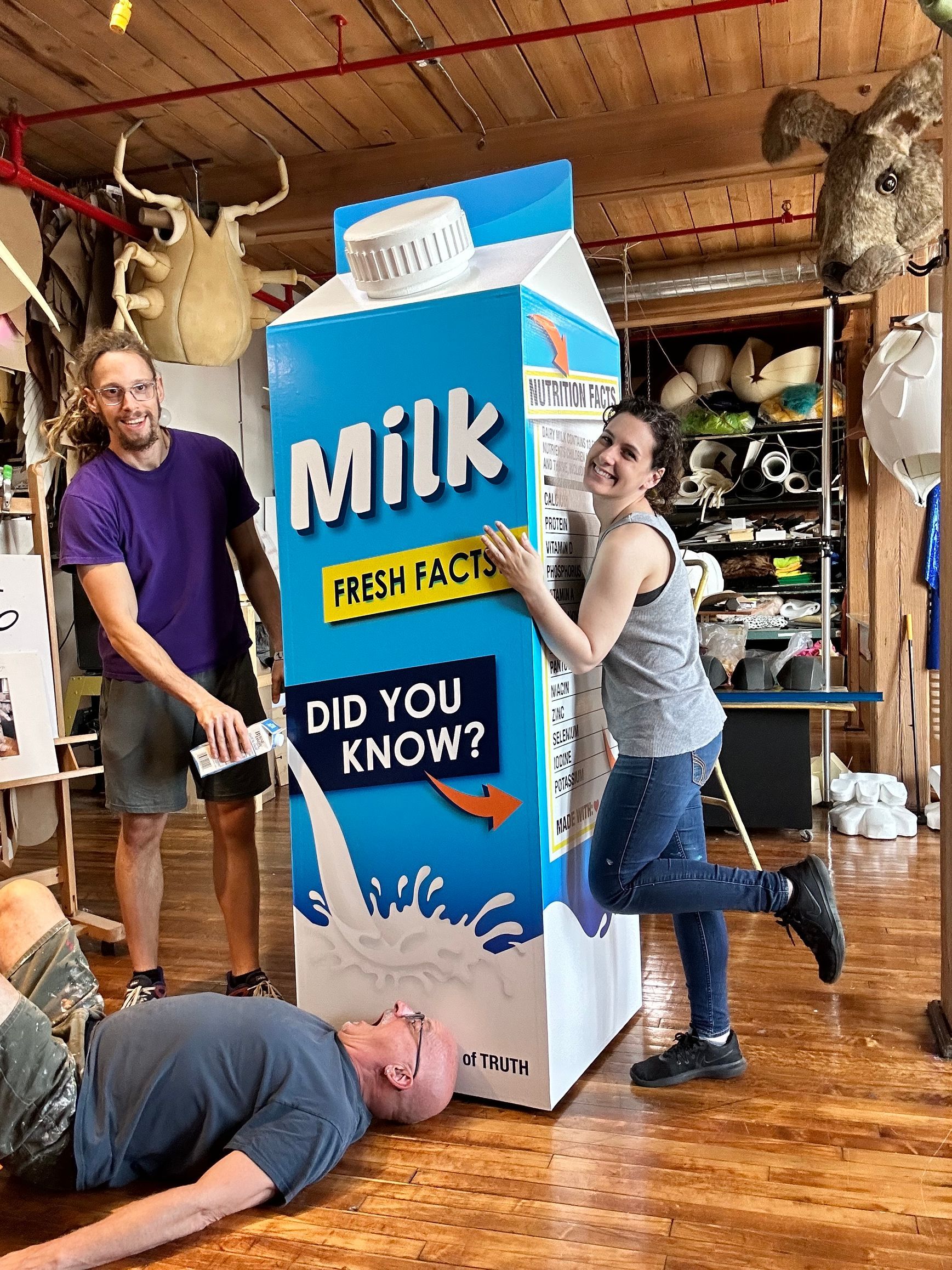Three people pose with a giant milk carton; one person pretends to be spilling out milk.