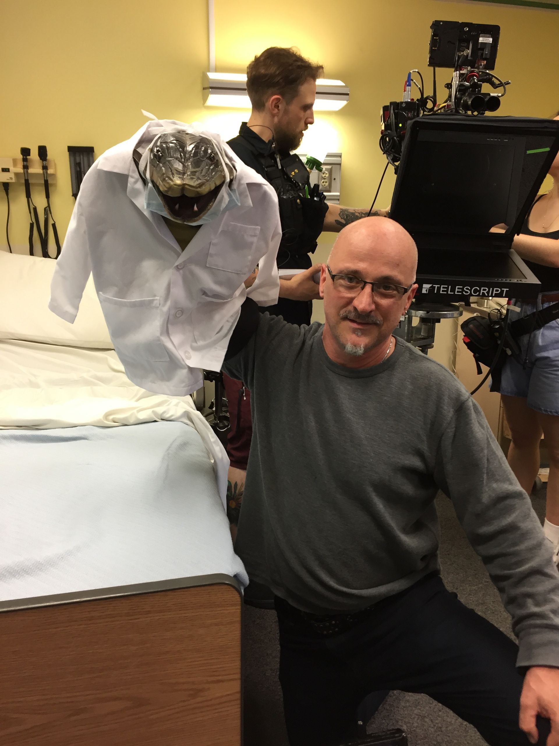 A bald man is holding a white shirt over a bed.