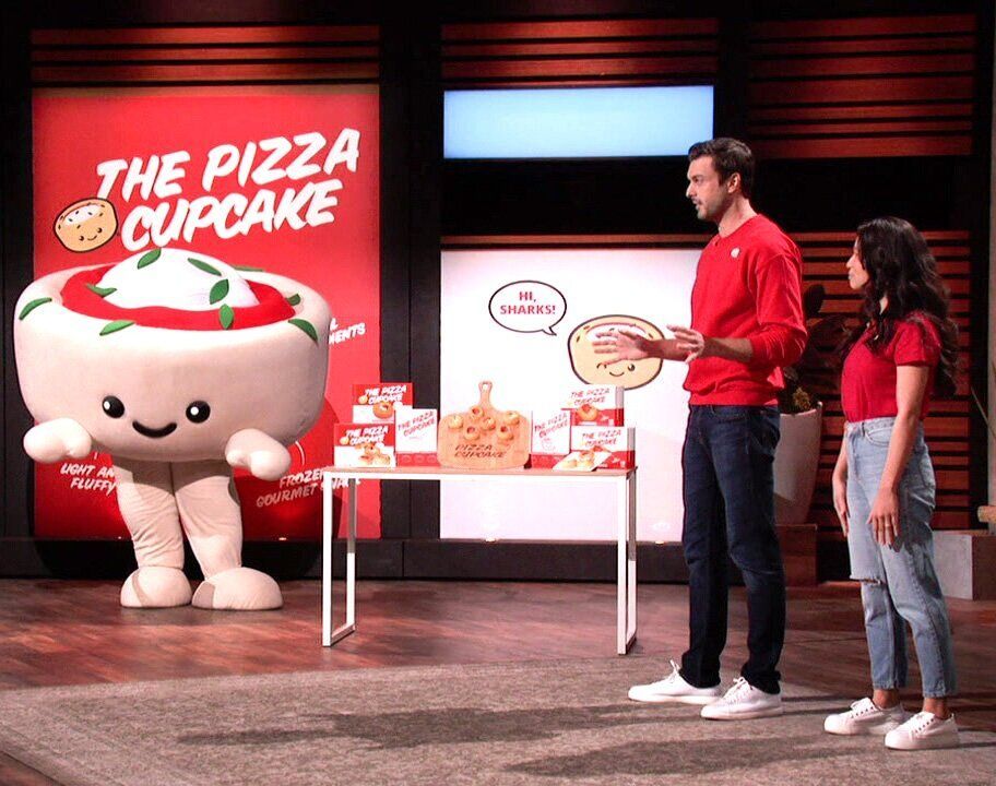 A man and a woman are standing in front of a pizza cupcake mascot.