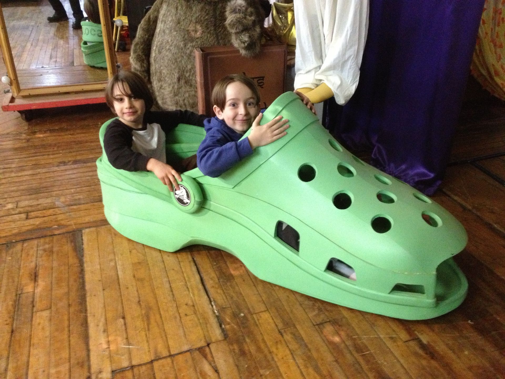 Two children are sitting in a green shoe on a wooden floor.
