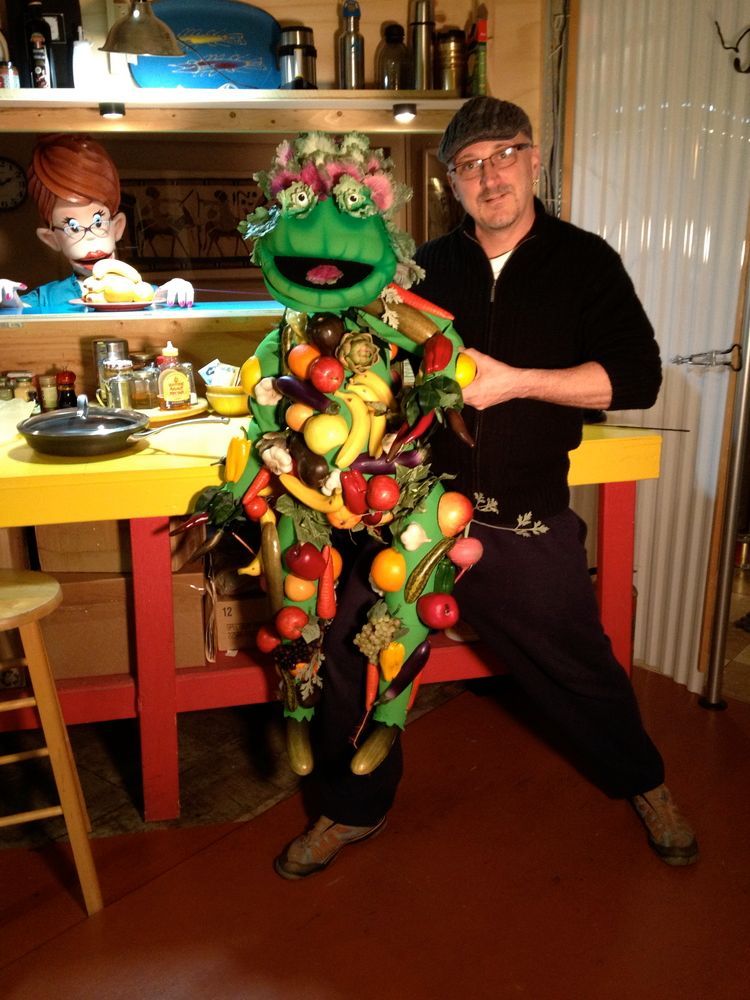 A man is kneeling next to a puppet made of fruits and vegetables