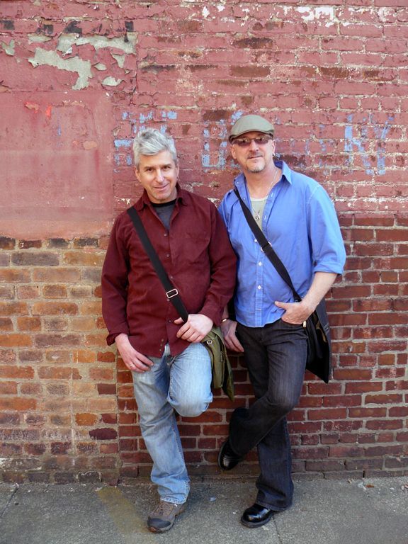Two men standing next to each other in front of a brick wall