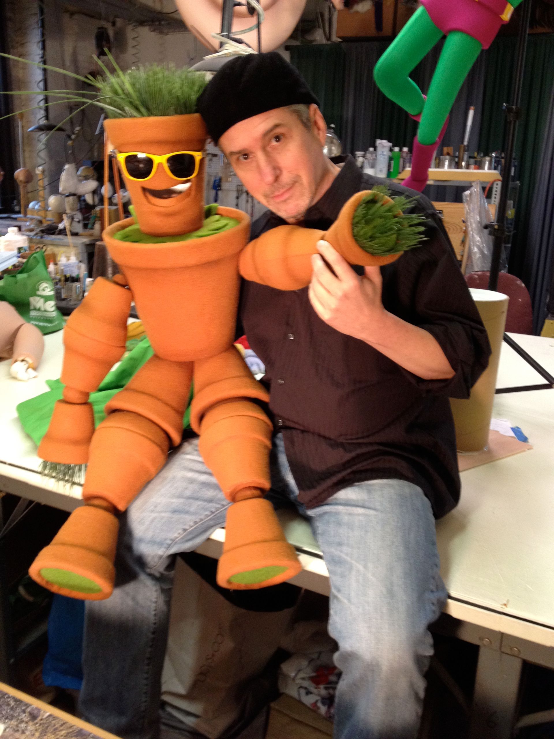 A man is sitting next to a stuffed carrot wearing sunglasses