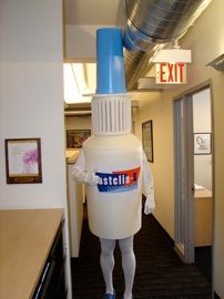 A person dressed as a bottle of glue is standing in a hallway.
