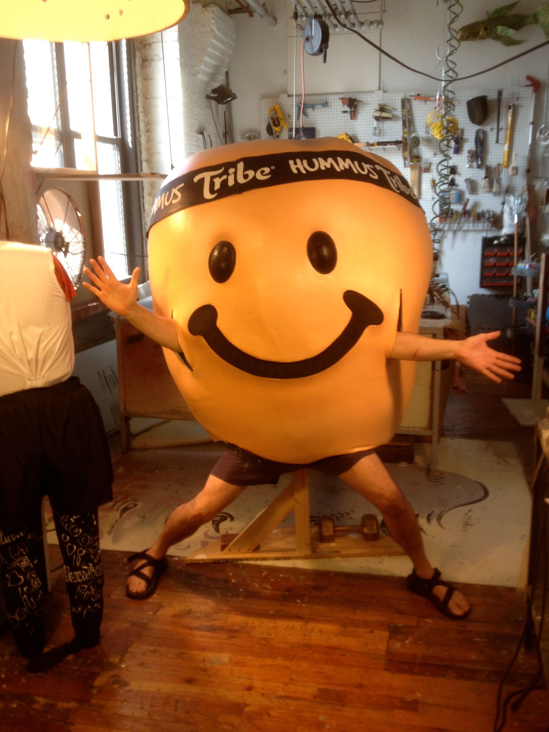 A man in a tribe hummus costume stands in a room