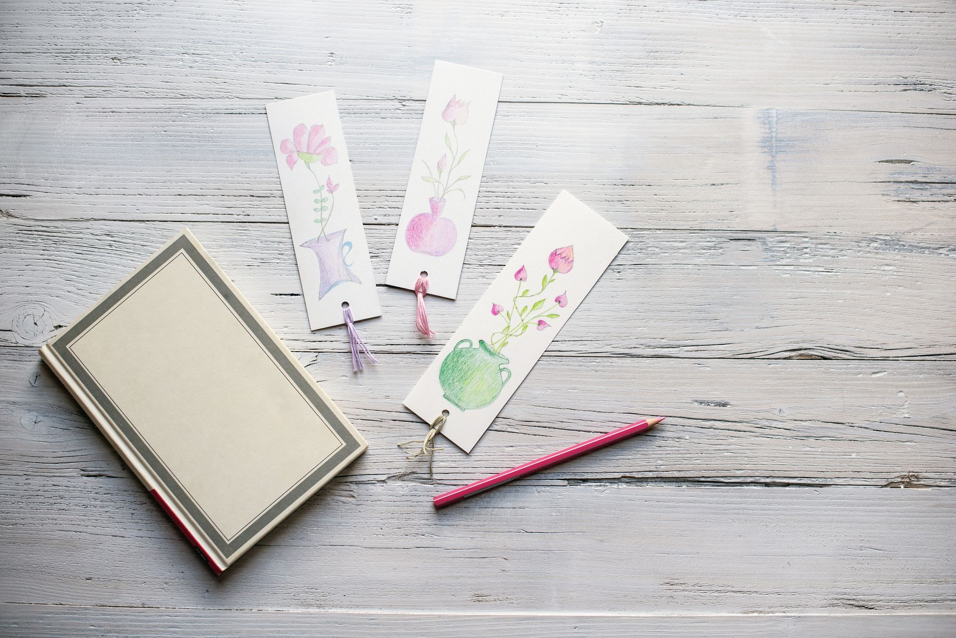 Three bookmarks are sitting on a wooden table next to a book and a pencil.