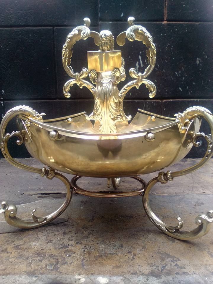 Cuenco ornamentado de latón dorado con asas con volutas y base decorativa. El cuenco presenta un elemento central detallado con detalles curvos.