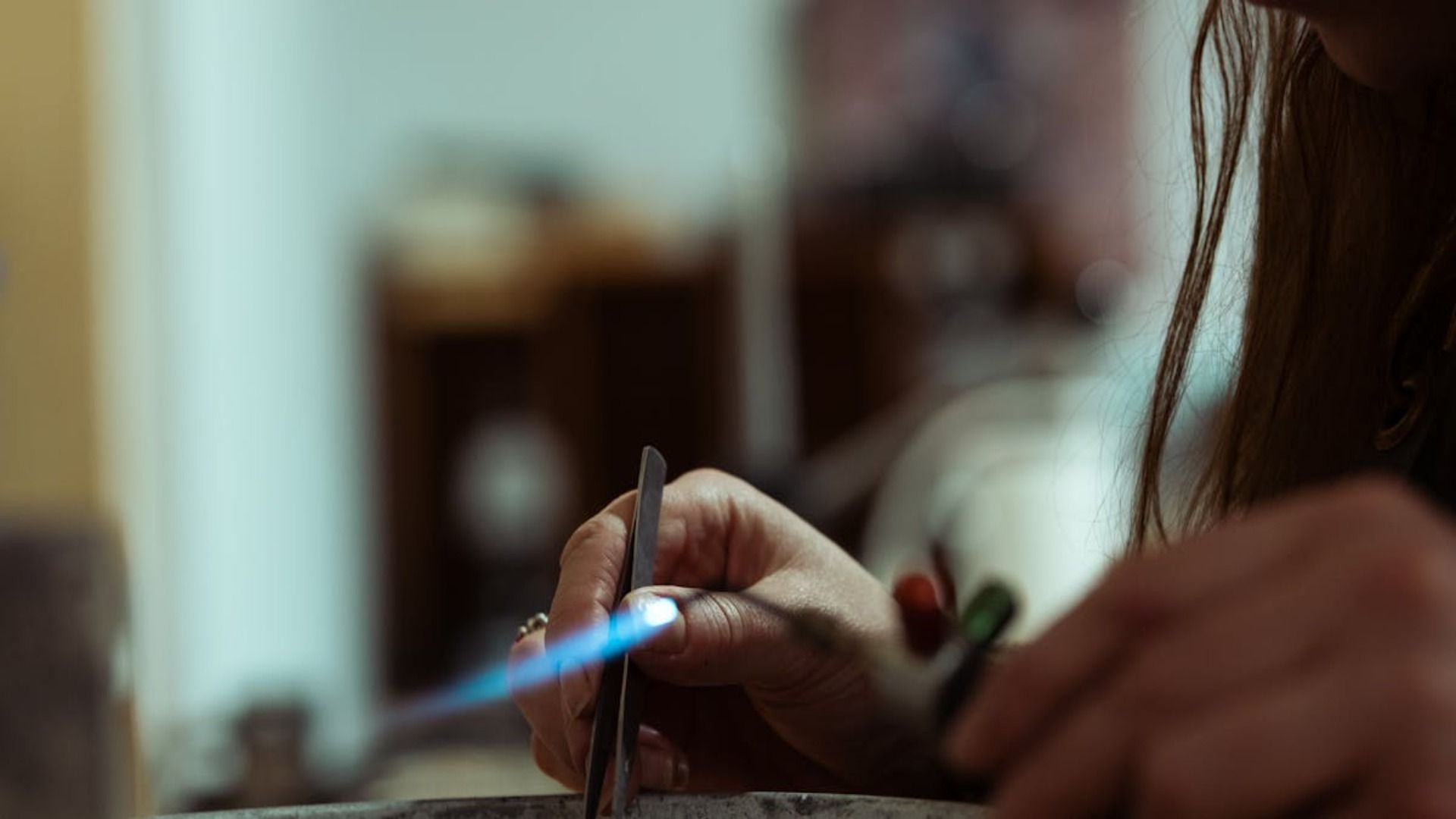 Las manos de una persona usando un soplete para calentar una pieza de metal, probablemente para hacer joyas. La llama es azul.