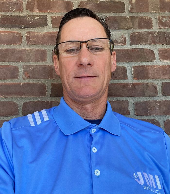 A man wearing glasses and a blue shirt is standing in front of a brick wall.