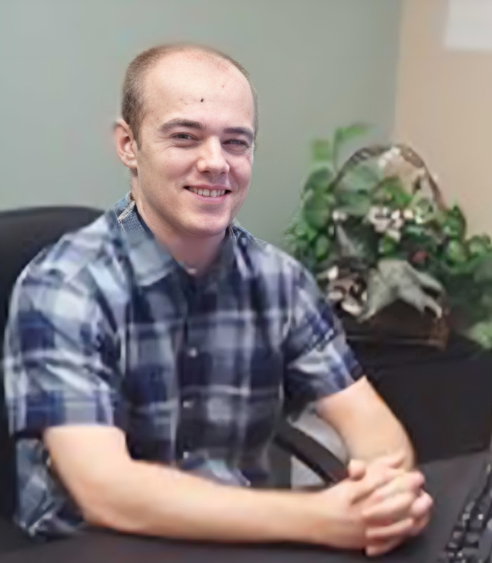 A man in a plaid shirt is sitting at a desk with his hands folded