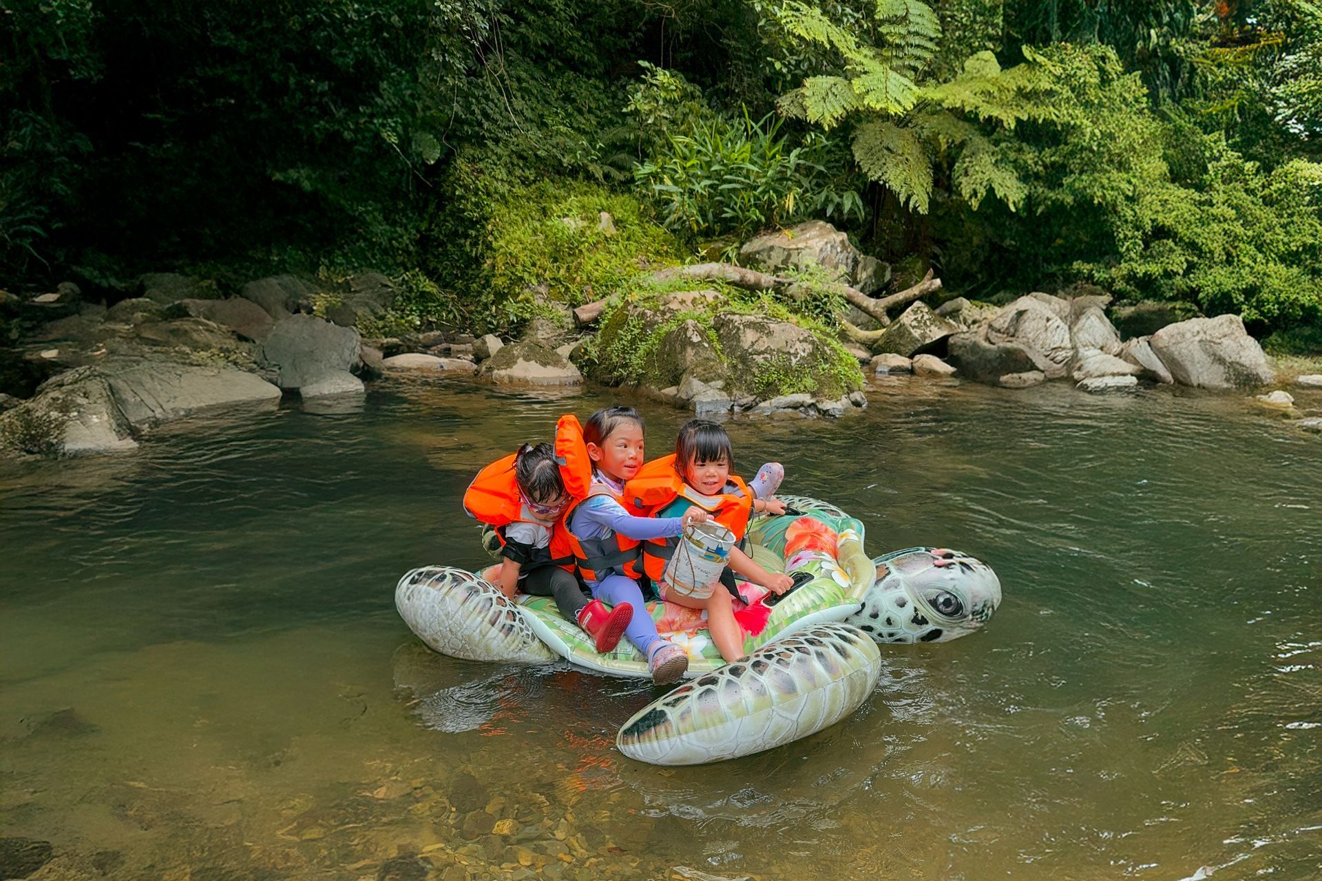 三個穿著救生衣的孩子乘坐烏龜形狀的充氣浮墊,在森林環繞的平靜多石的小溪上漂浮。