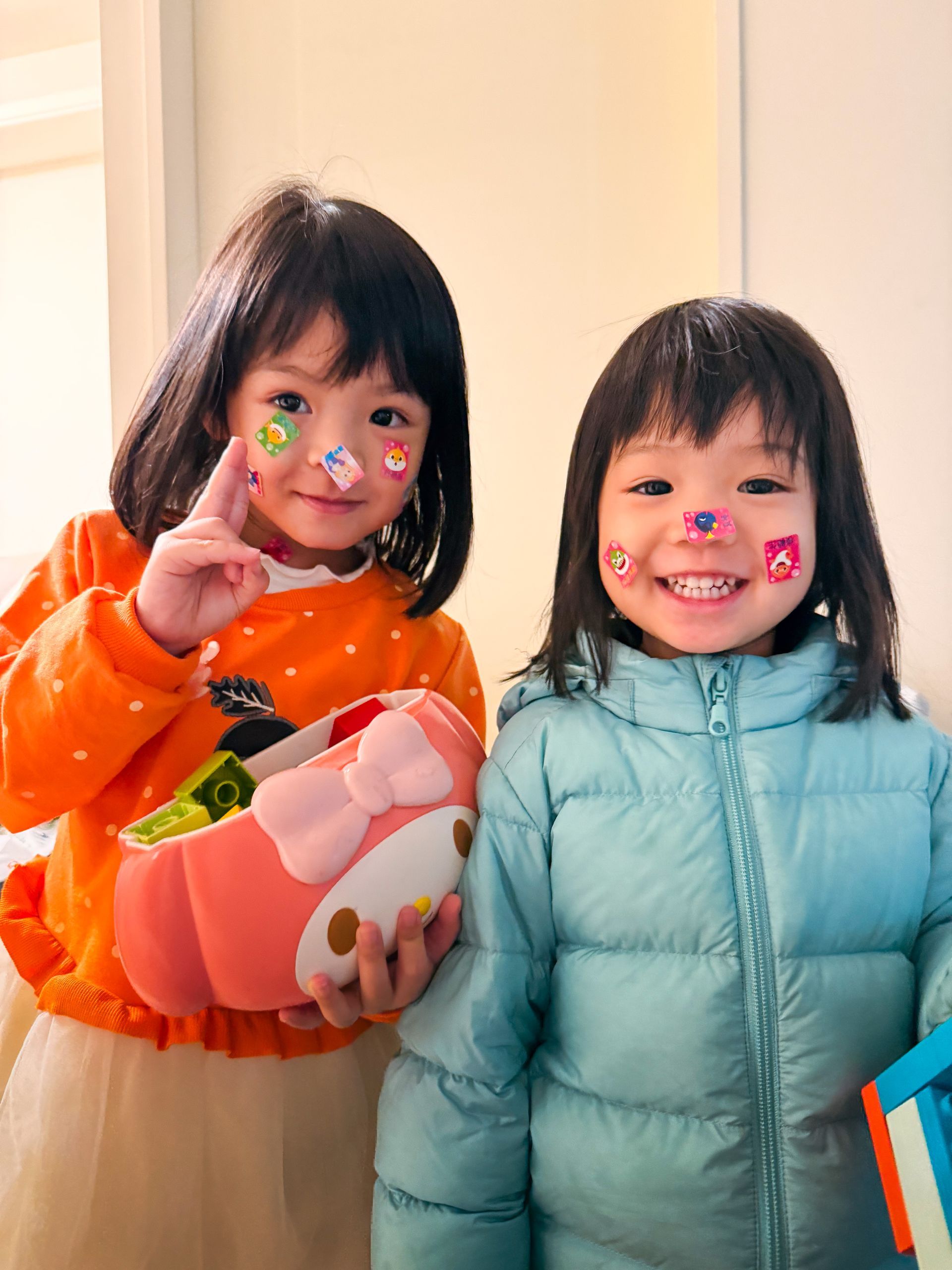 兩個臉上貼著貼紙的孩子麵帶微笑,一個穿著橘色上衣,拿著粉紅色書包,另一個穿著藍色外套。