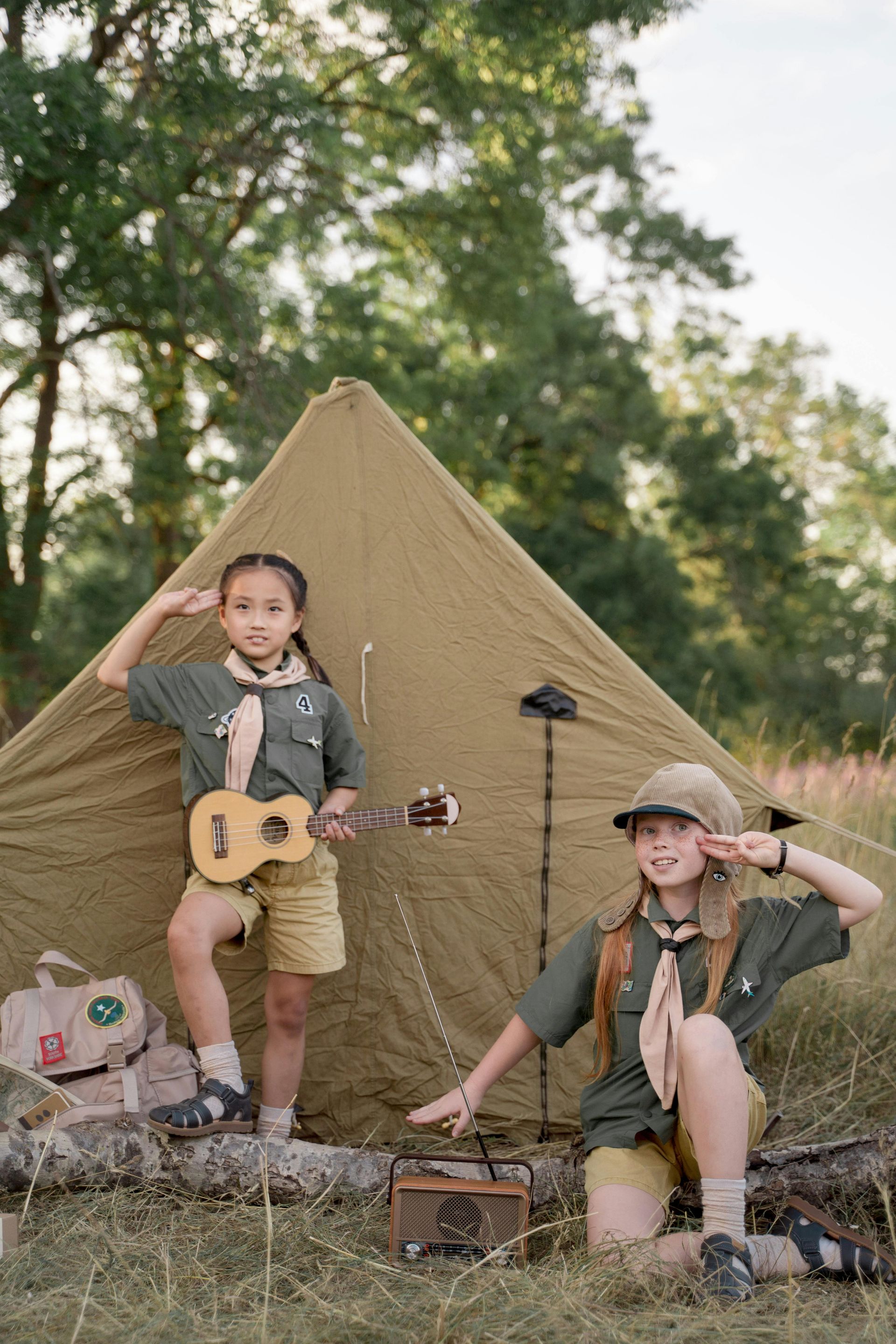 兩個身穿童子軍制服、頭戴童子軍帽的孩子在田野裡的帳篷旁擺姿勢拍照,其中一個拿著尤克里裡,兩人都敬禮。