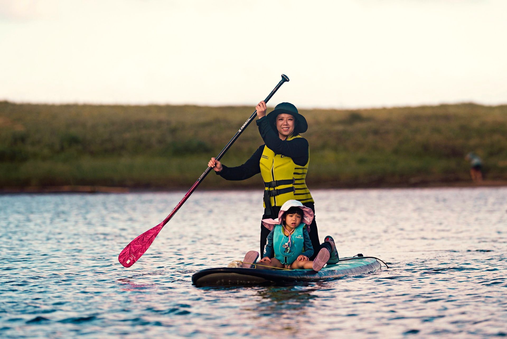 一名成人和一名兒童穿著救生衣在平靜的水面上劃槳板，背景是綠草茵茵的岸邊。
