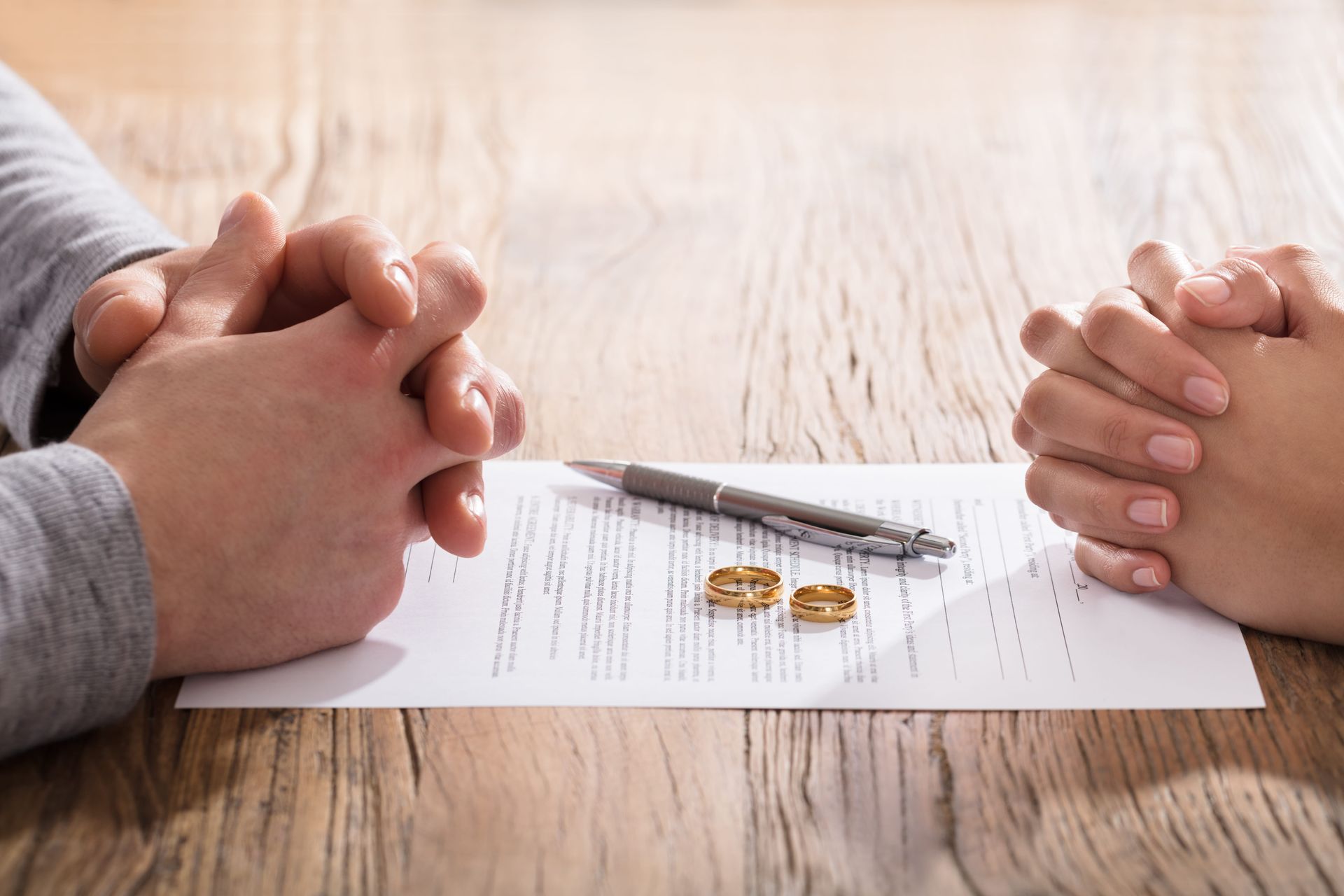Two pairs of hands rest on a table beside a document, a pen, and two gold wedding rings, suggesting a divorce process.