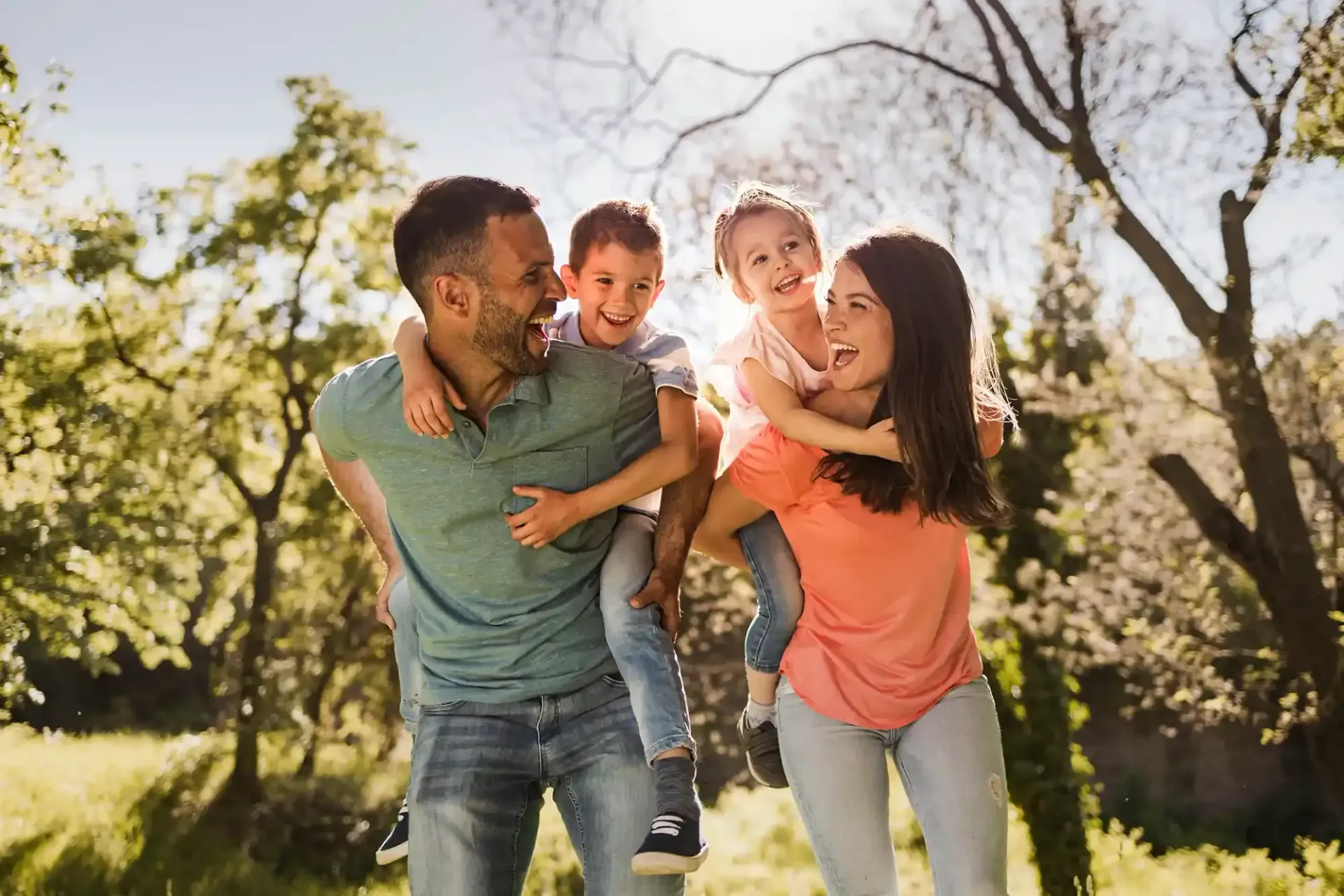 Parents giving children piggyback rides in a sunny park; laughing, joyful expressions.