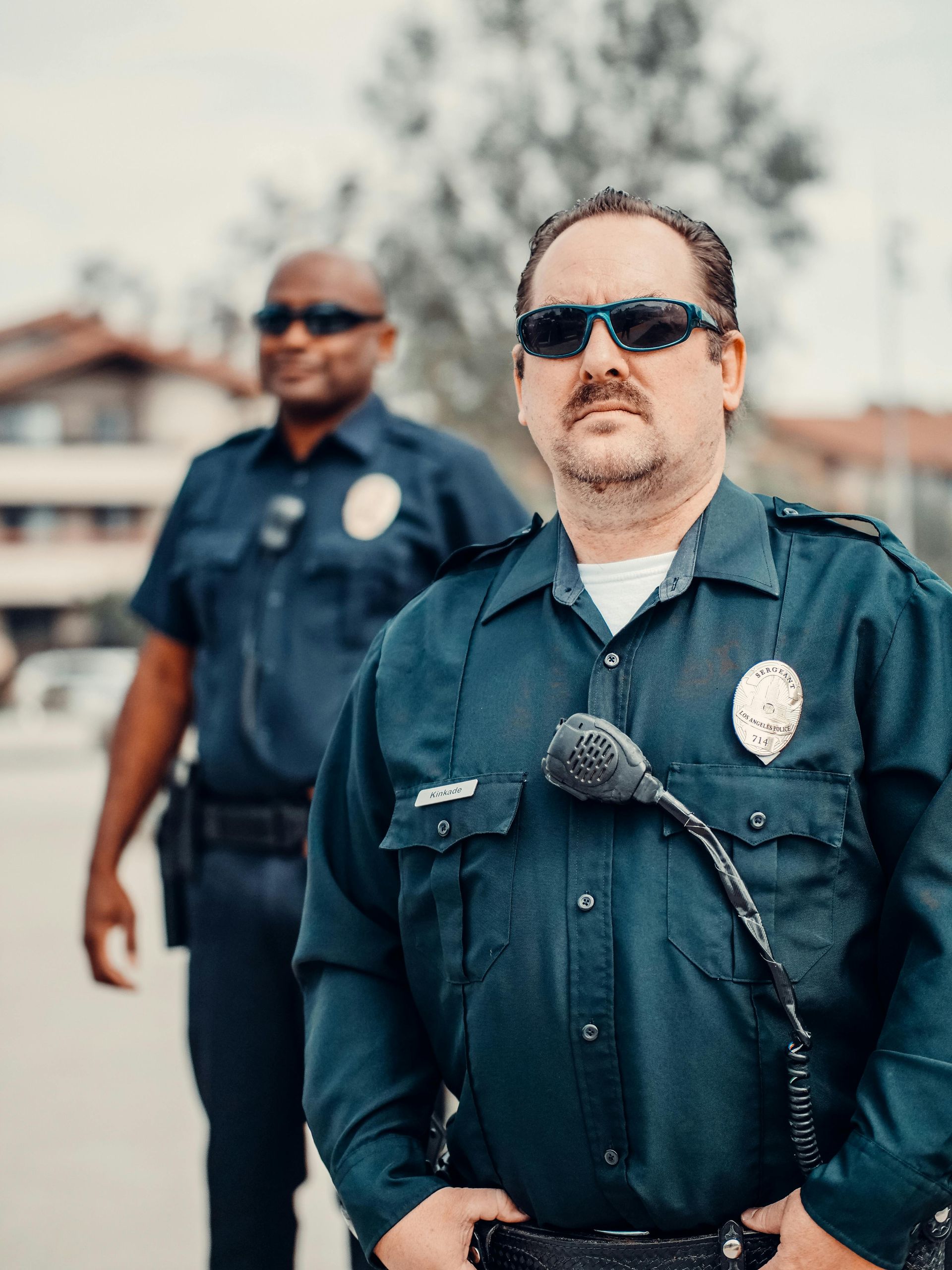Two police officers in uniform, outdoors. One stands in front, other blurred in background. Both wear sunglasses.