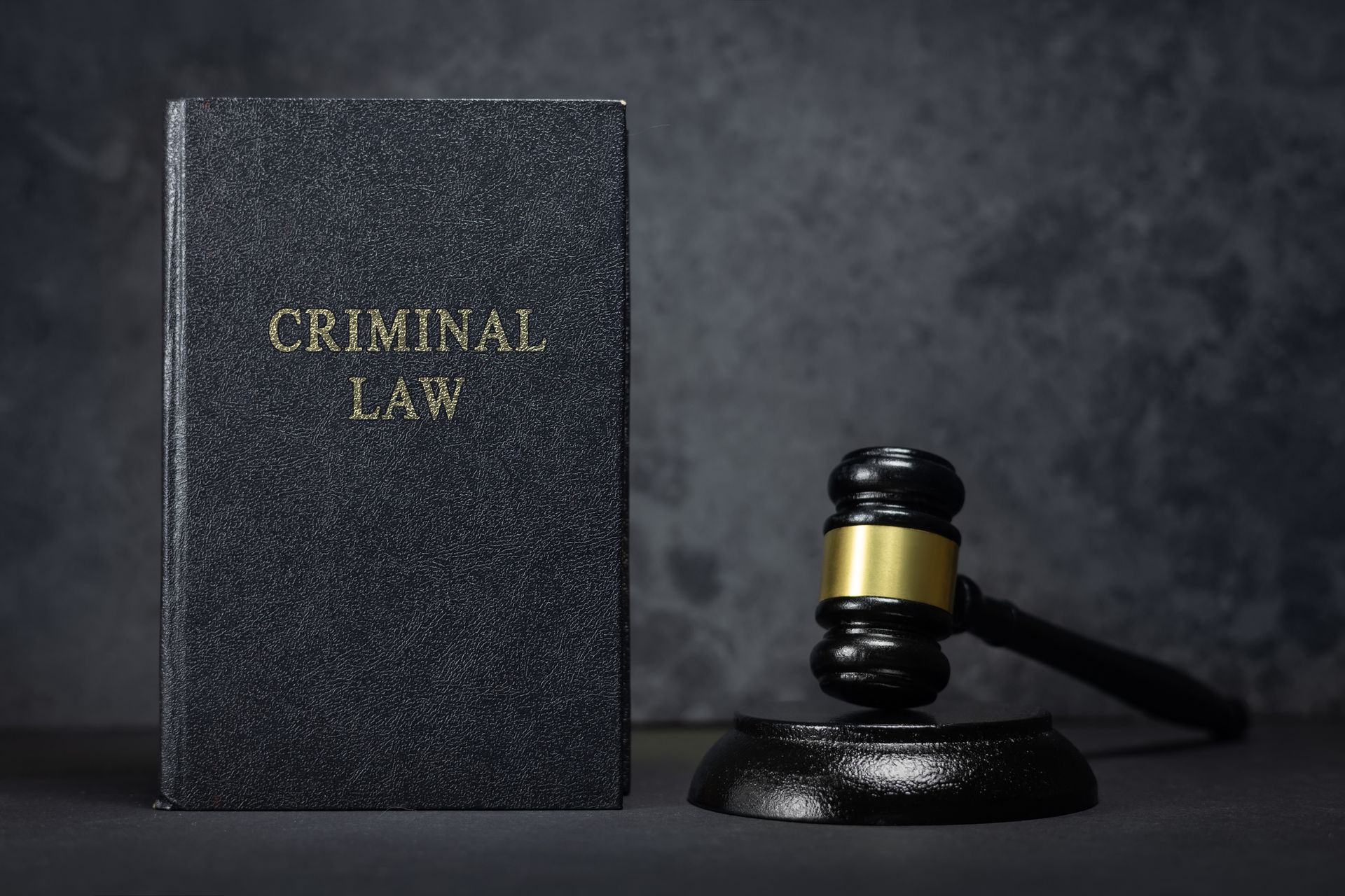 Black criminal law book standing next to a wooden judge’s gavel on a dark surface.