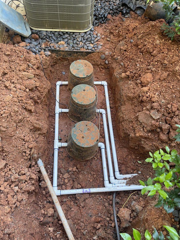 Three concrete lids connected by white PVC pipes in an excavated trench. A shovel leans nearby.