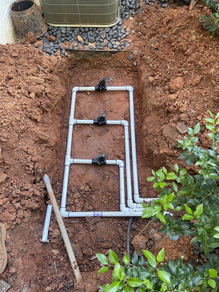Trench in dirt with three irrigation valves and PVC pipes, near an AC unit and plants.