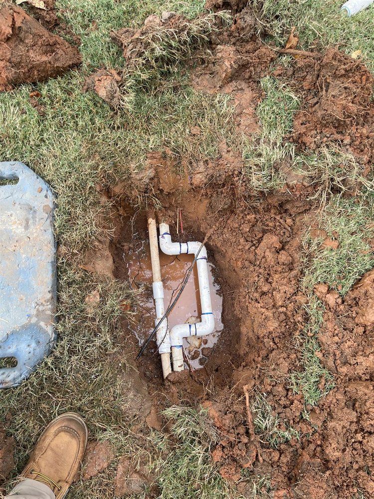 Pipes in a small excavated hole in grassy ground, likely plumbing repair. Brown shoes on the bottom left.