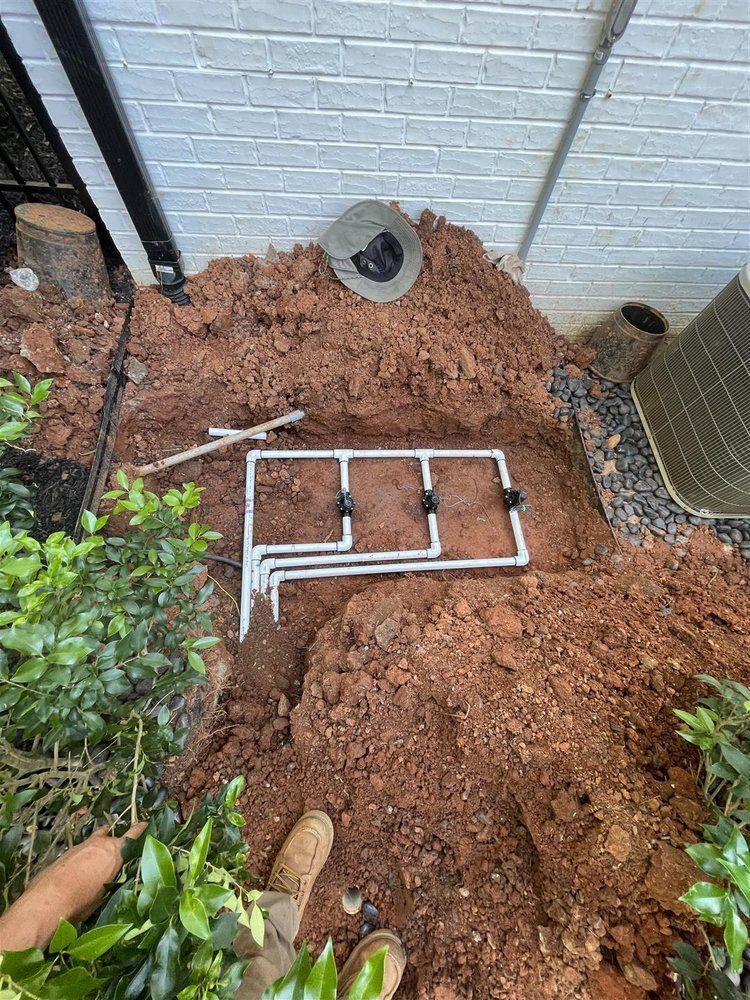A network of white PVC pipes with black connectors in a dug-up area of red soil, near a building.