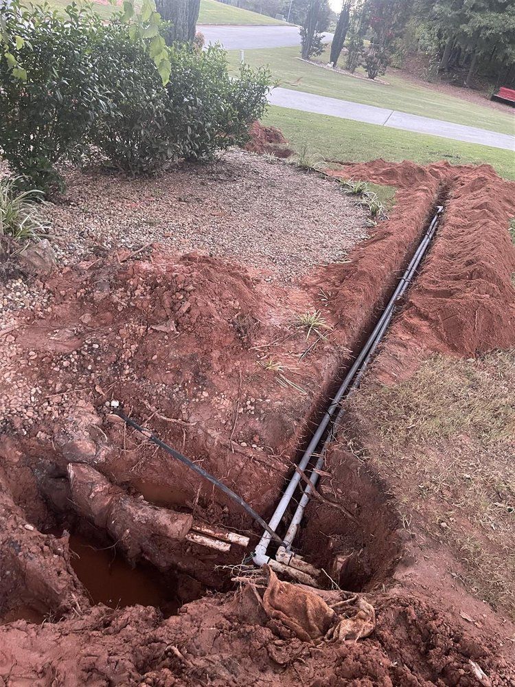 Trench dug in red dirt with pipes laid inside for a landscaping project near a curb and bush.